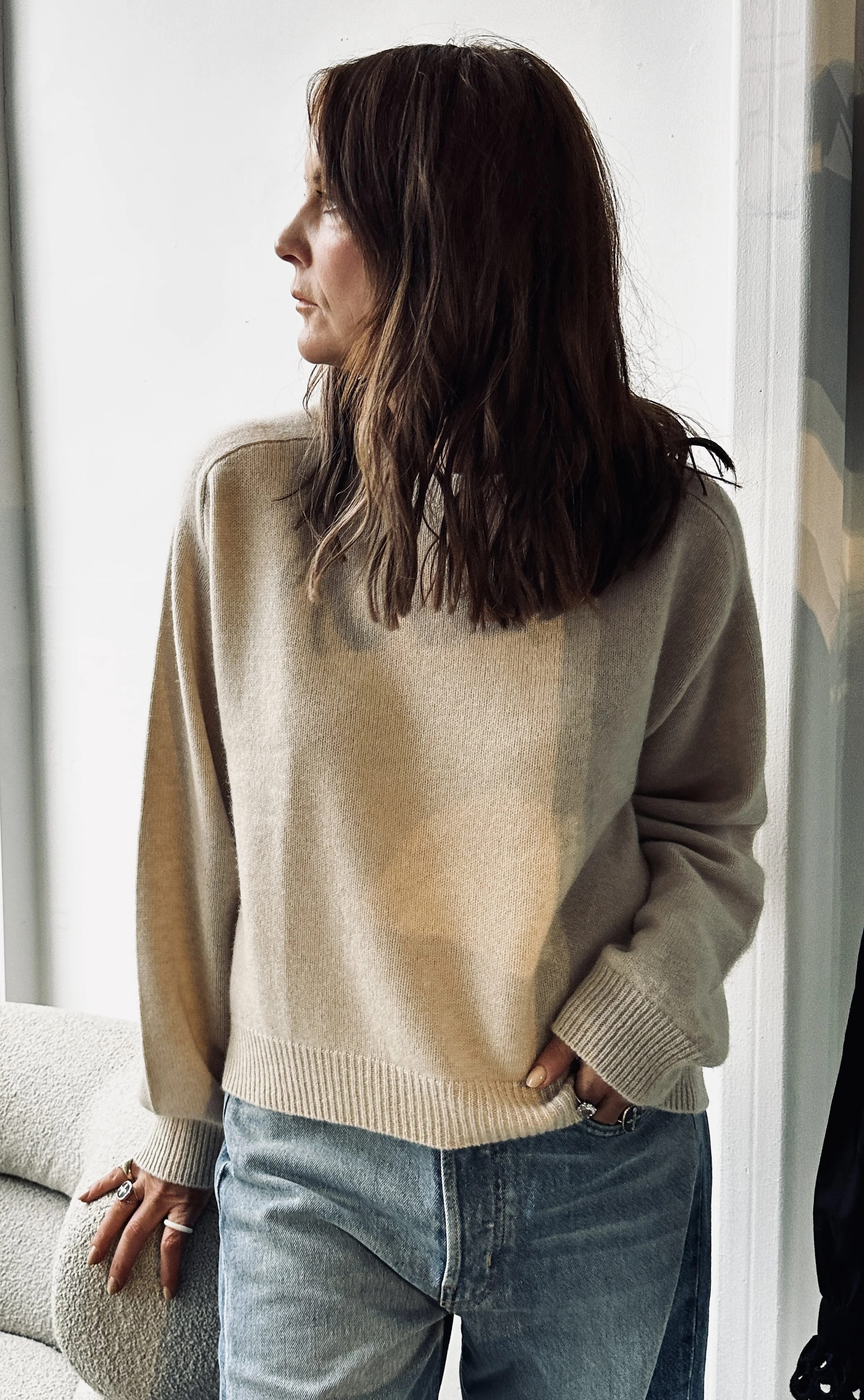 A woman with dark brown, wavy hair stands indoors next to a beige sofa. She wears a beige sweater and light blue jeans, and has rings on her fingers. She is looking to the left with one hand in her pocket.