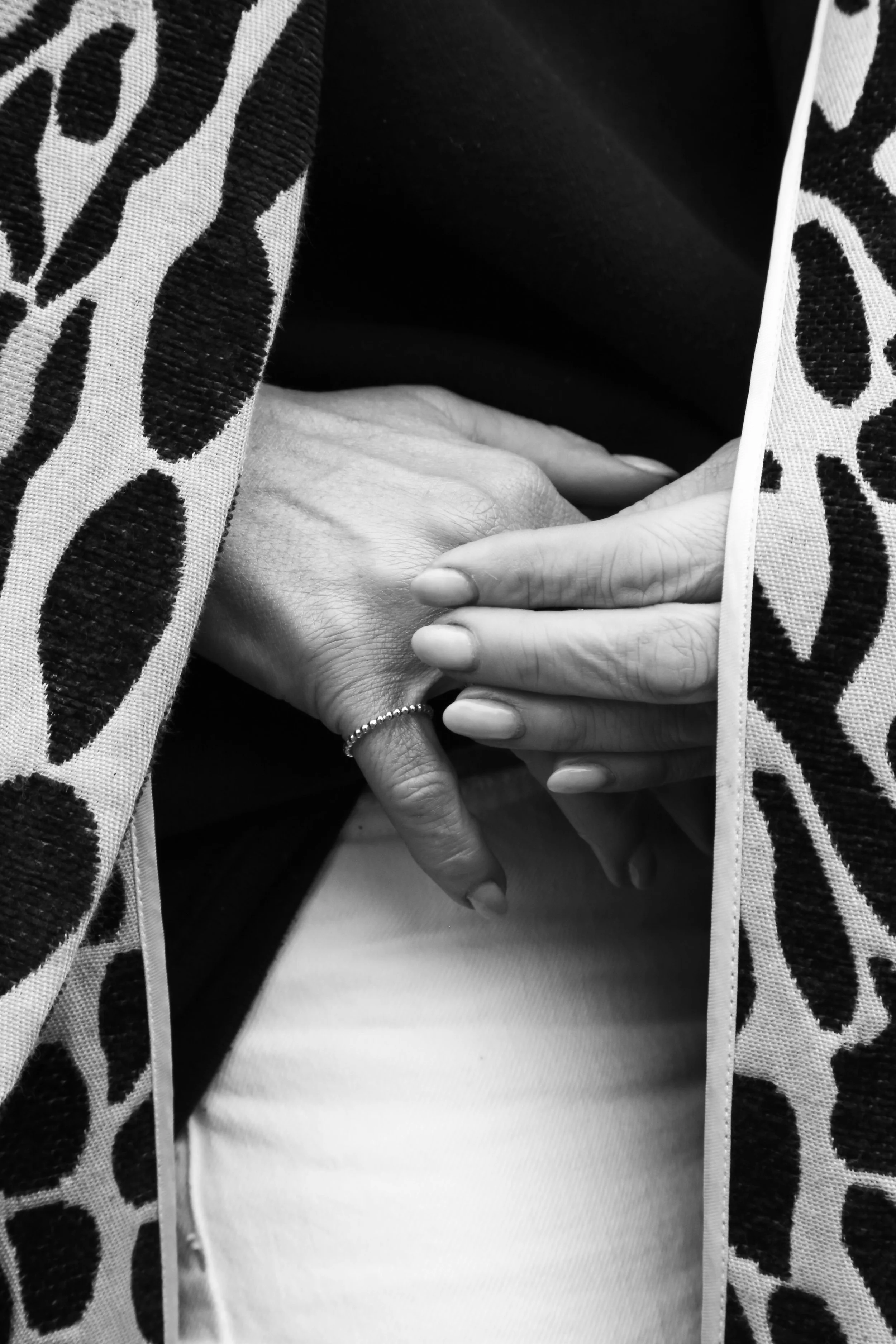 Black and white photograph of two hands gently clasped, one with a beaded ring and the other with neatly manicured nails, partially covered by a patterned textile fabric.