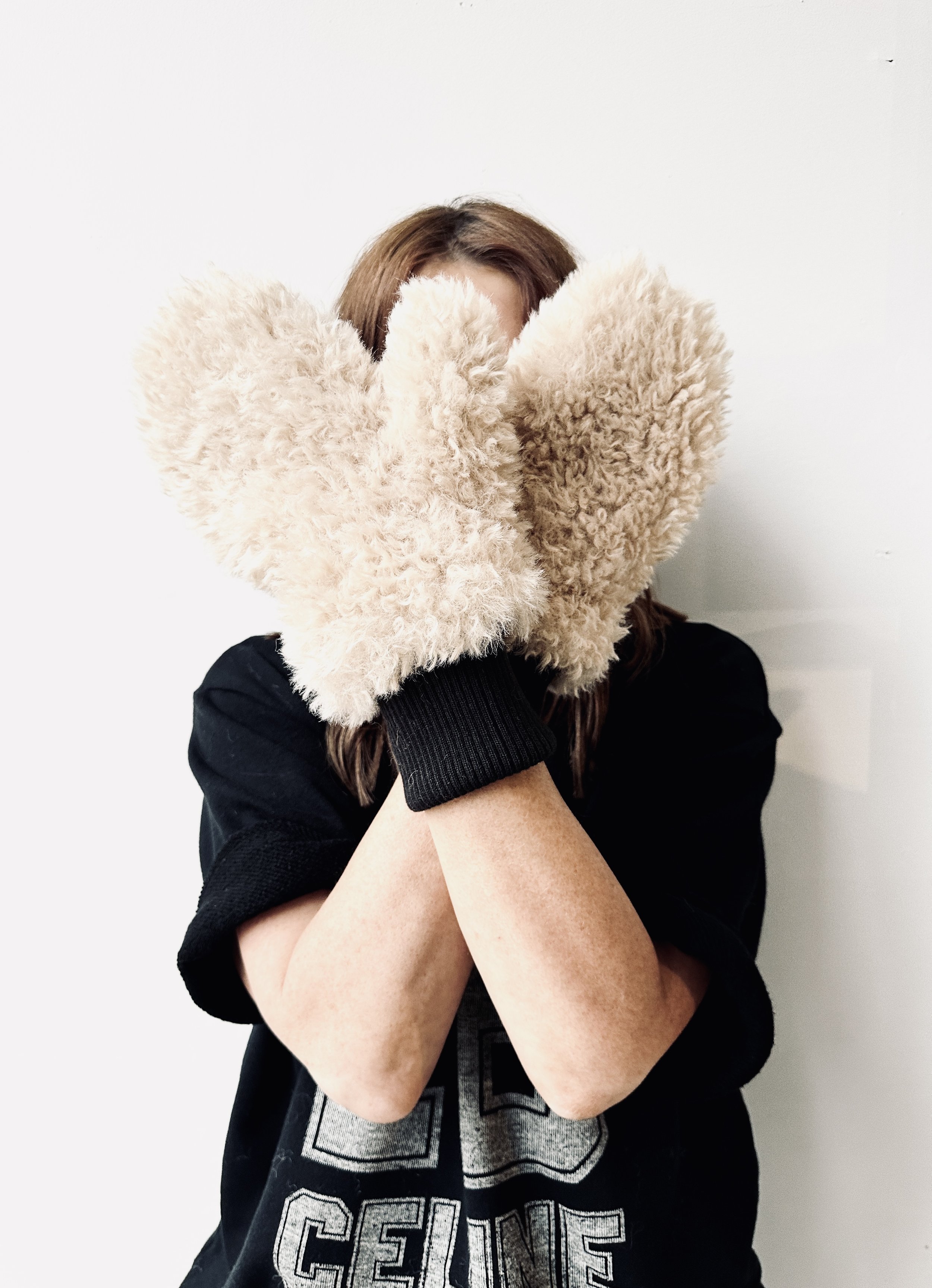 Person wearing a black T-shirt with a graphic on it, holding large beige fluffy mittens up to cover their face, standing against a white wall.