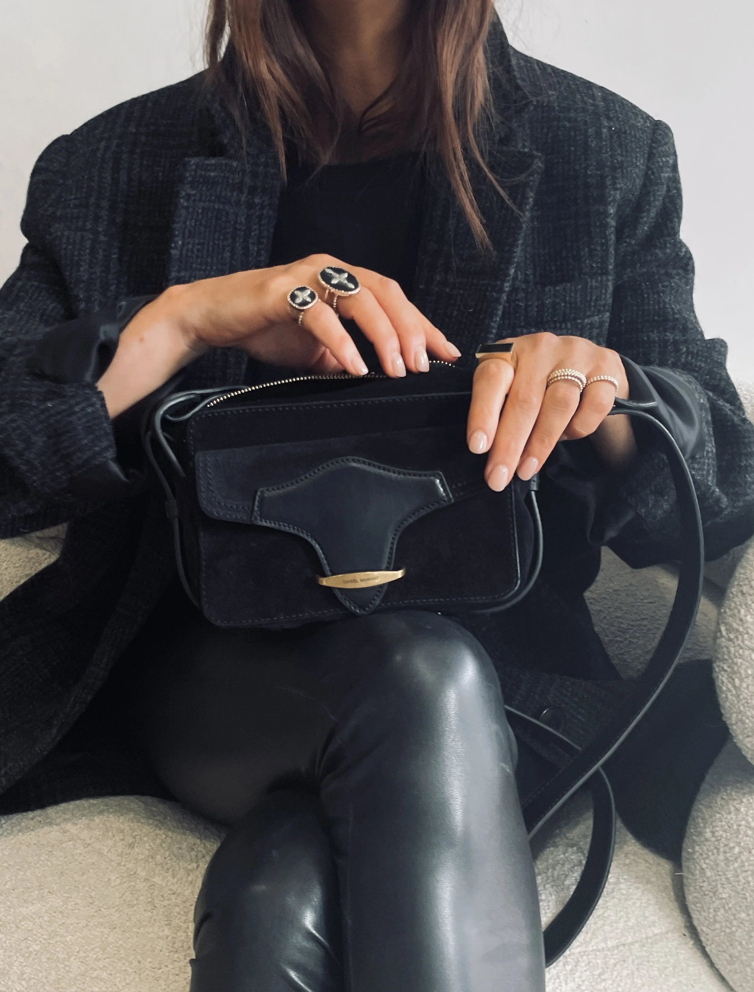 Person dressed in black and gray, holding a black designer handbag with gold accents, sitting on a neutral-colored seat.
