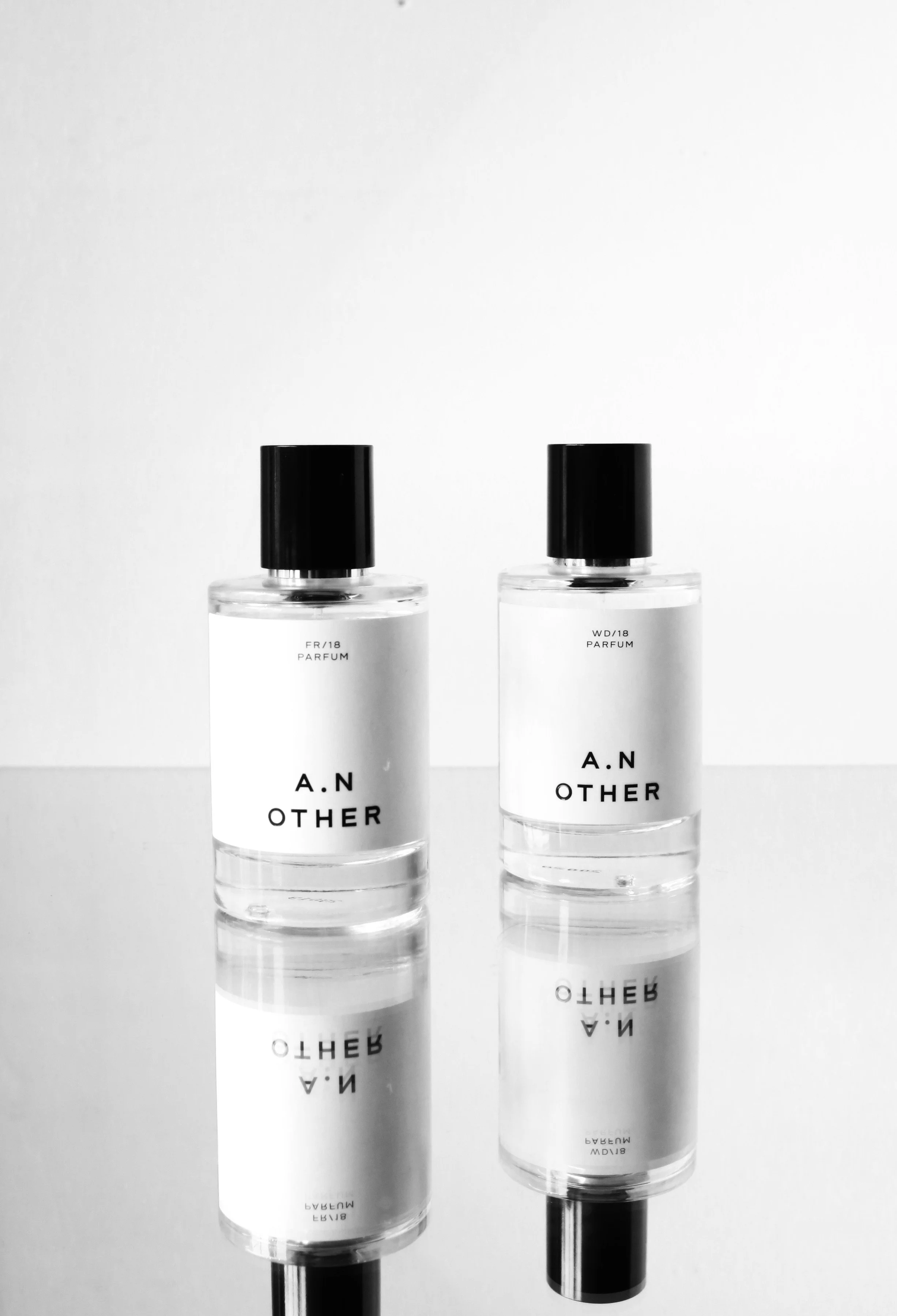 Two clear perfume bottles with black caps, labeled 'A.N OTHER,' are placed on a reflective surface against a plain white background.