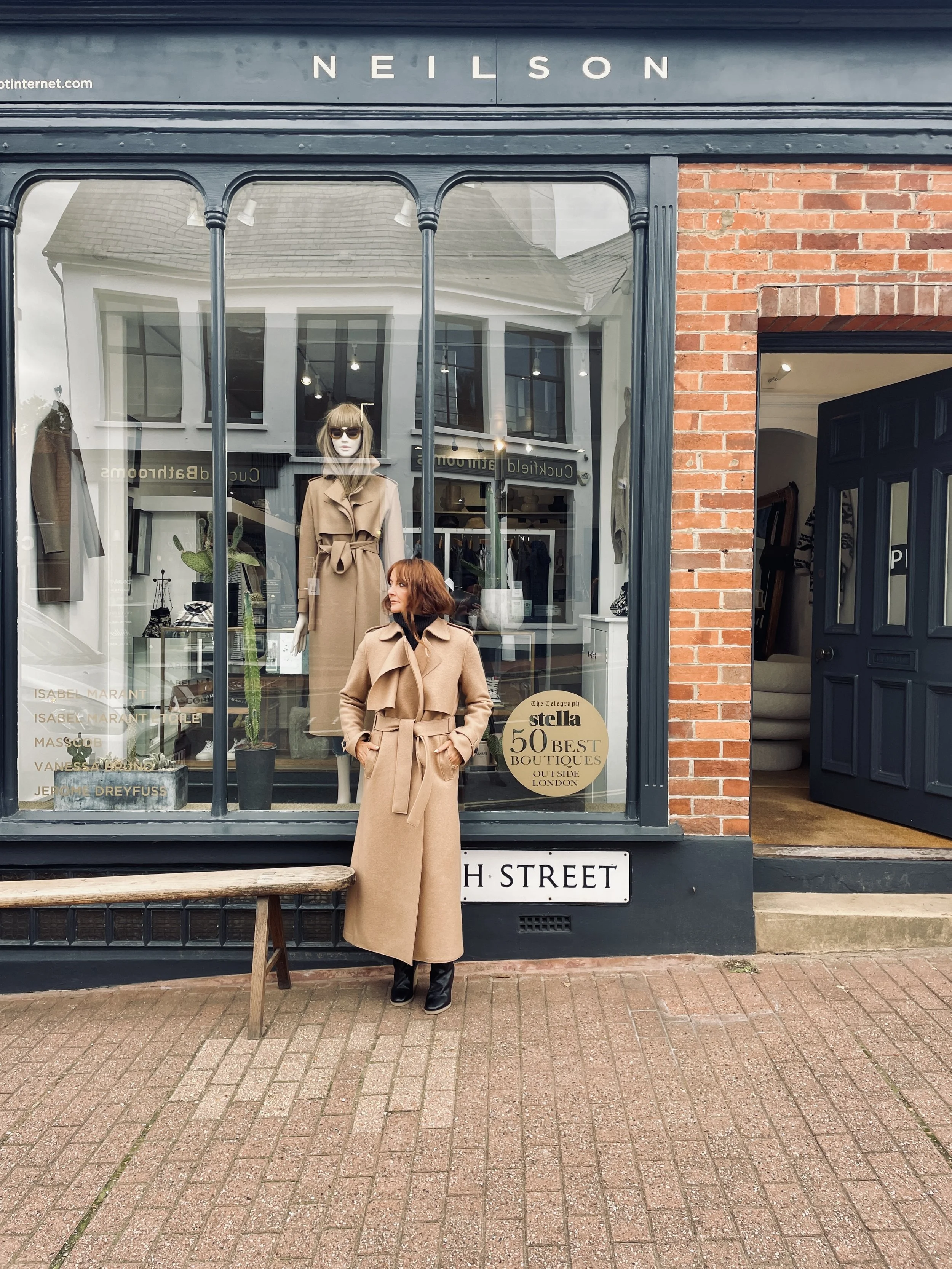 A woman with red hair wearing a beige coat standing outside a boutique store on High Street. The store window displays mannequins dressed in fashionable clothing and has signs recognizing it among the "50 Best Boutiques Outside London." The store has