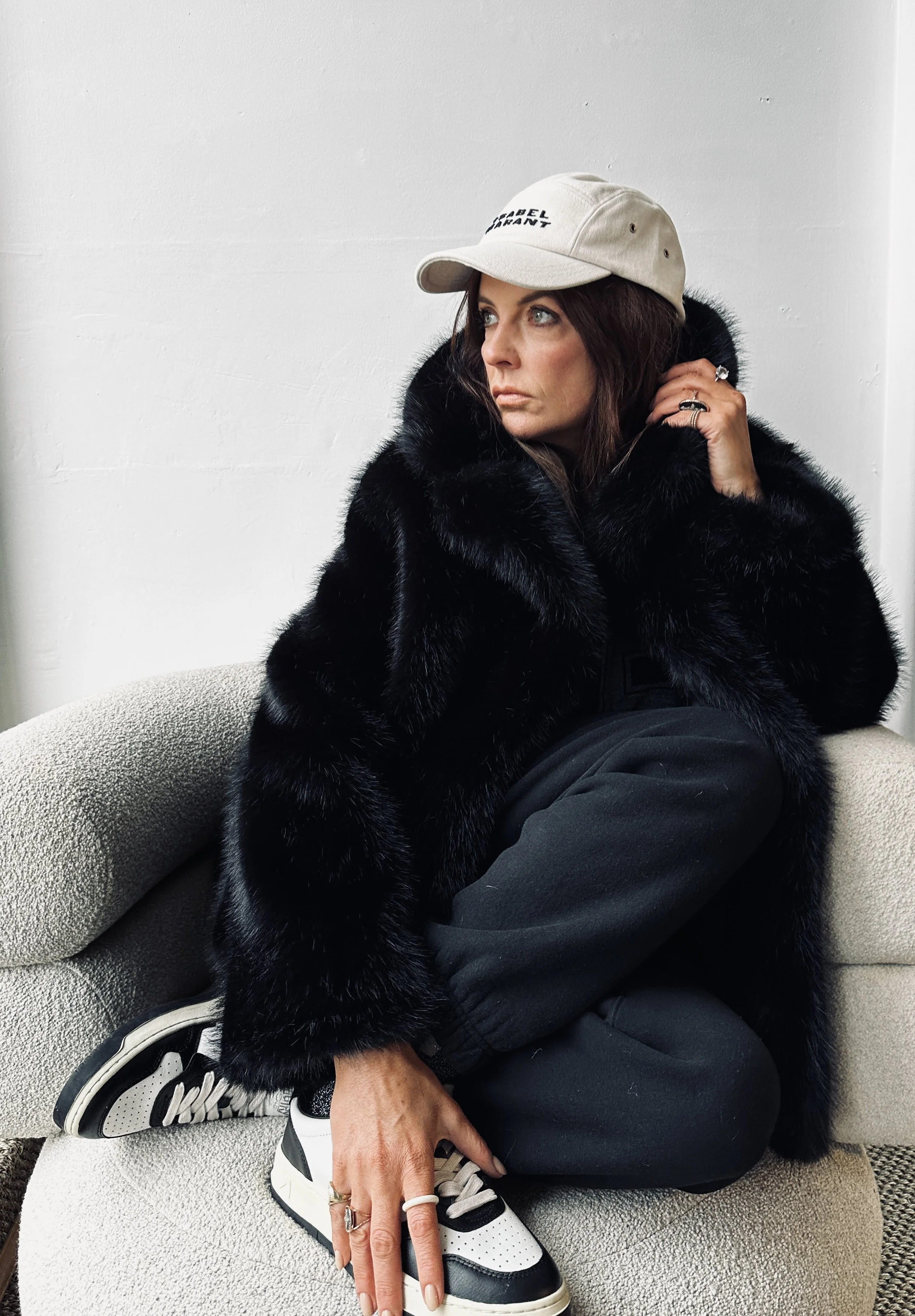 A woman with brown hair sitting on a beige couch, wearing a black fur coat, black pants, white and black sneakers, and a beige baseball cap, looking thoughtfully to the side with her right hand touching her hair.