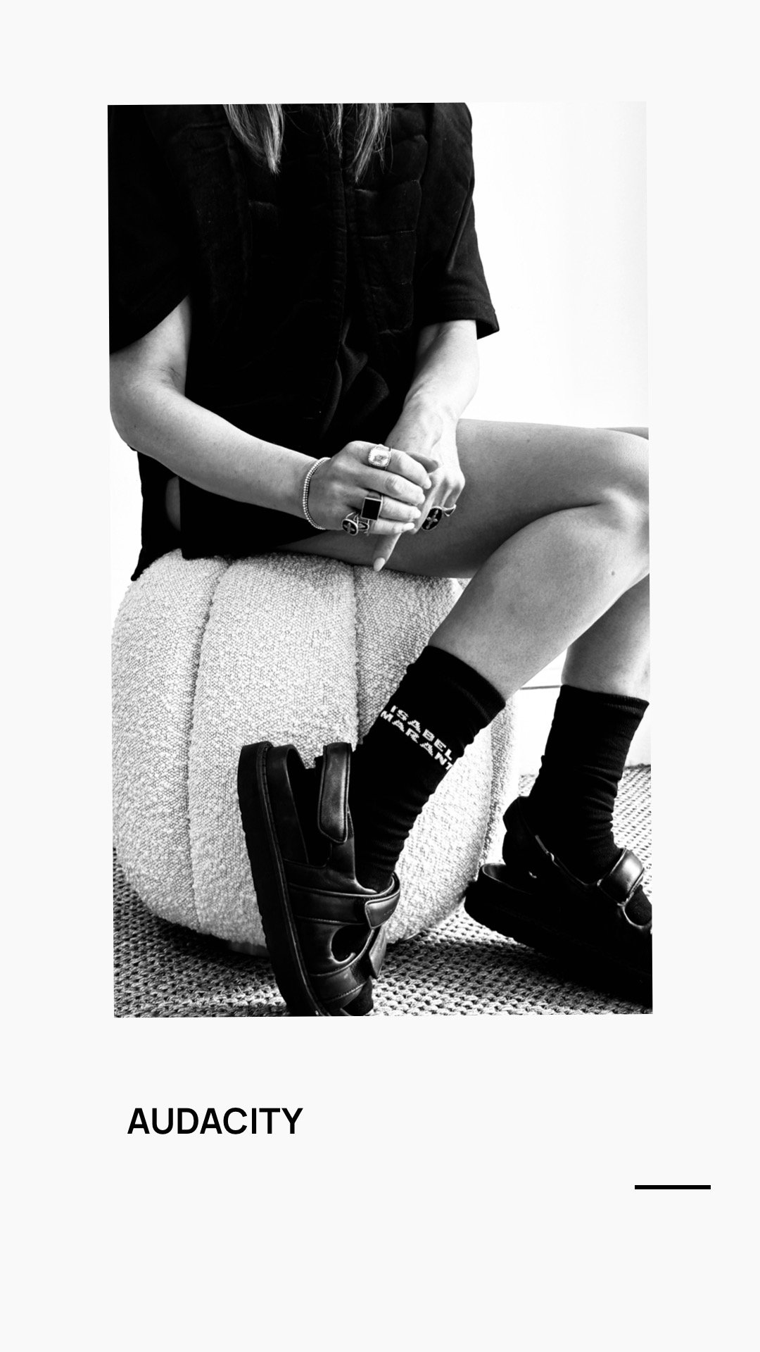Black and white photo of a person sitting on a textured ottoman, wearing black sandals, black socks with white text, rings, and a black shirt, with sitting posture and jewelry visible.