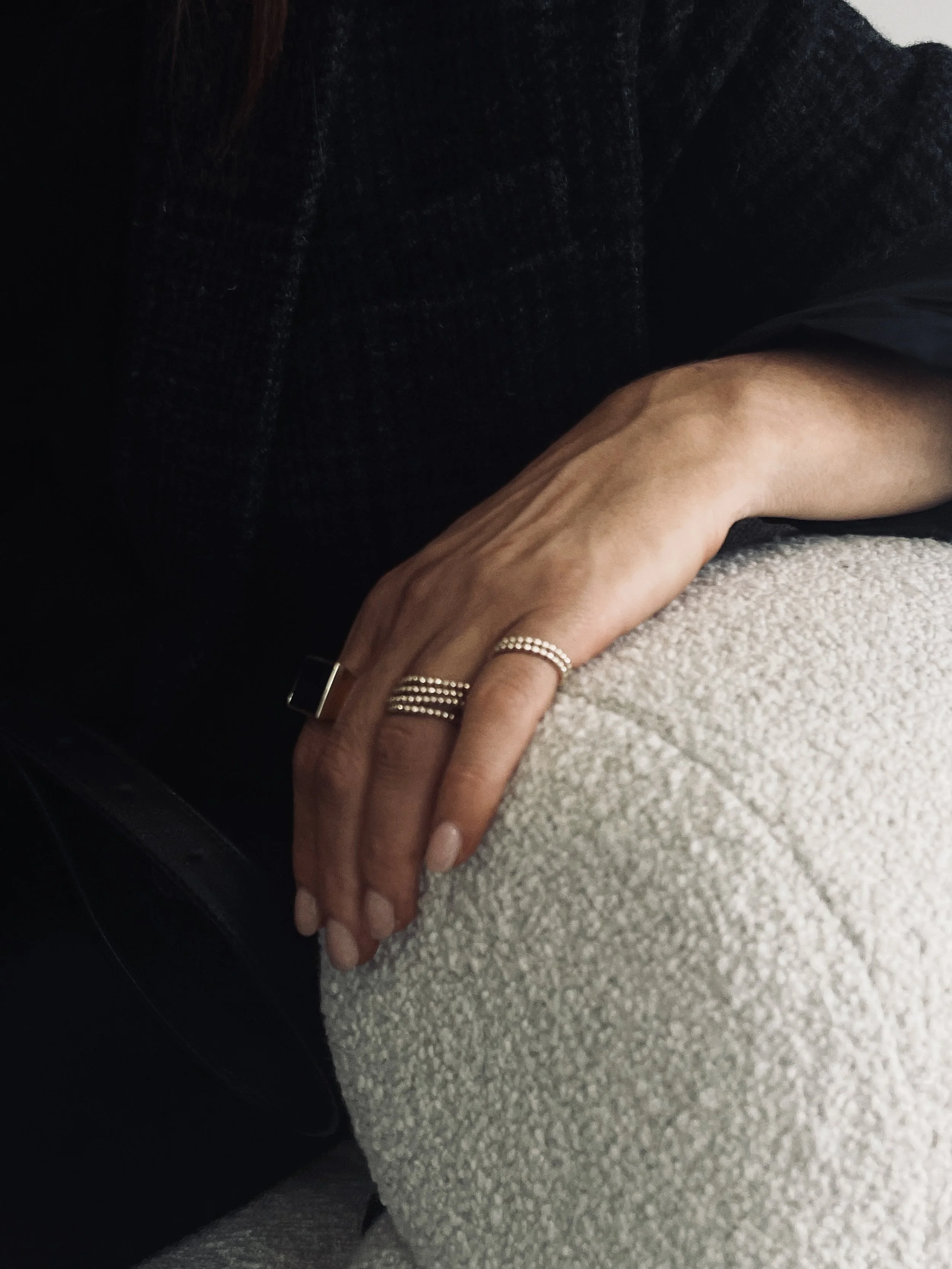 Close-up of a person's hand resting on a person's knee, wearing rings and jewelry, with a dark top and light-colored textured fabric.