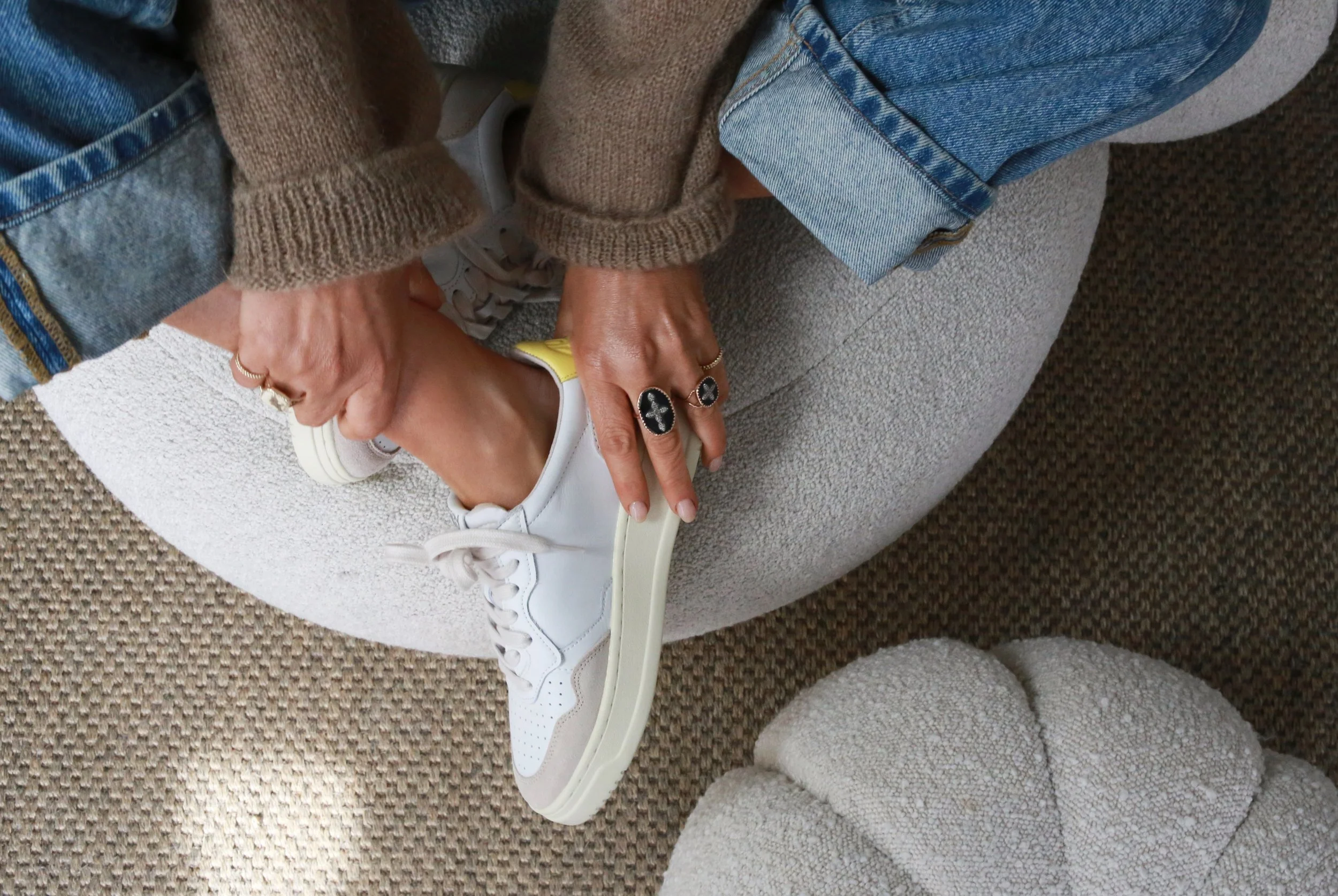 Person sitting on a light-colored rounded couch, tying the laces of their white sneakers with yellow accents. They are wearing blue jeans and a brown sweater, with multiple rings on their fingers. The setting includes textured, neutral-toned cushions