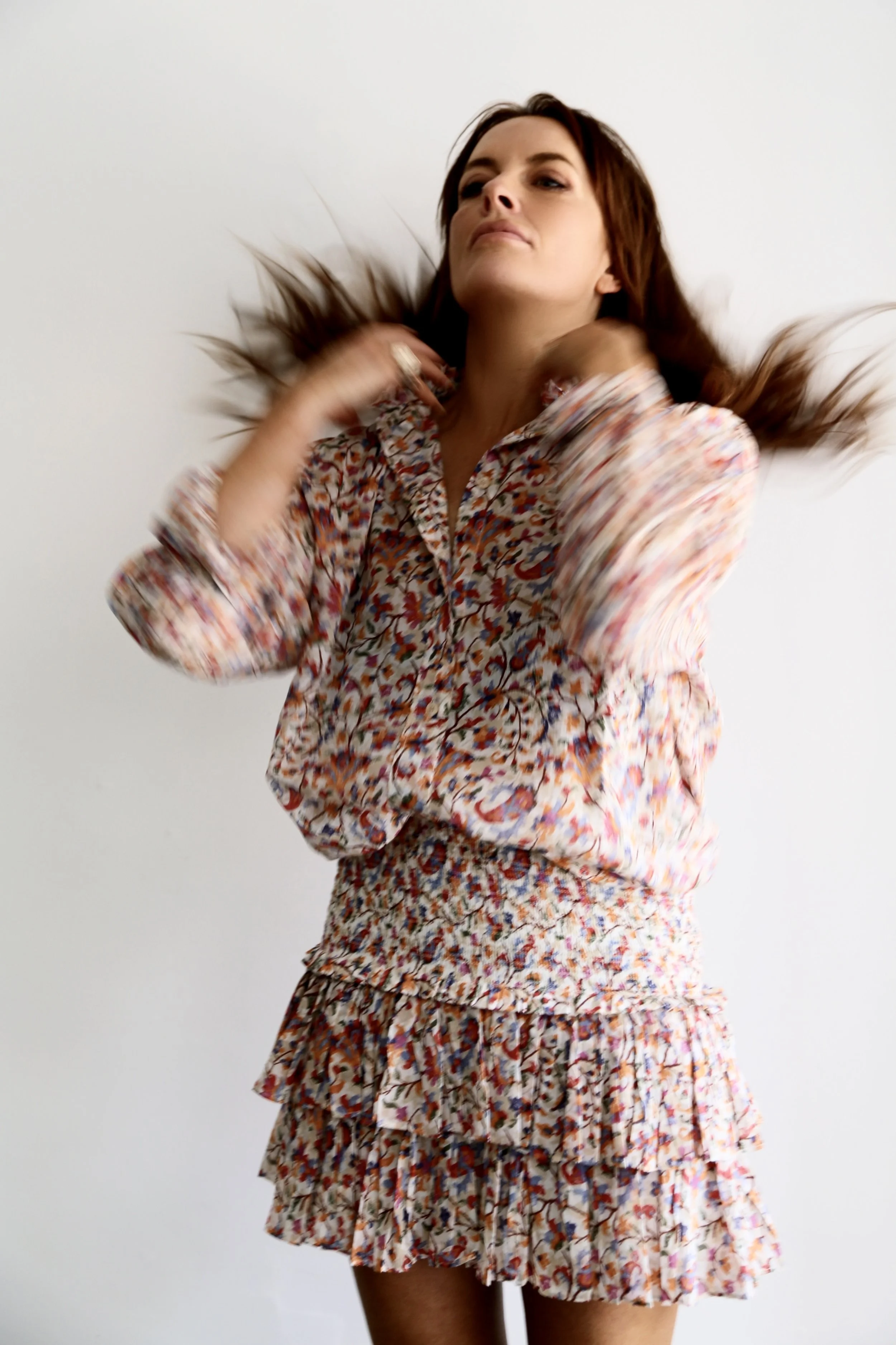 A woman with reddish-brown hair wearing a floral patterned dress, flipping her hair against a plain white background.
