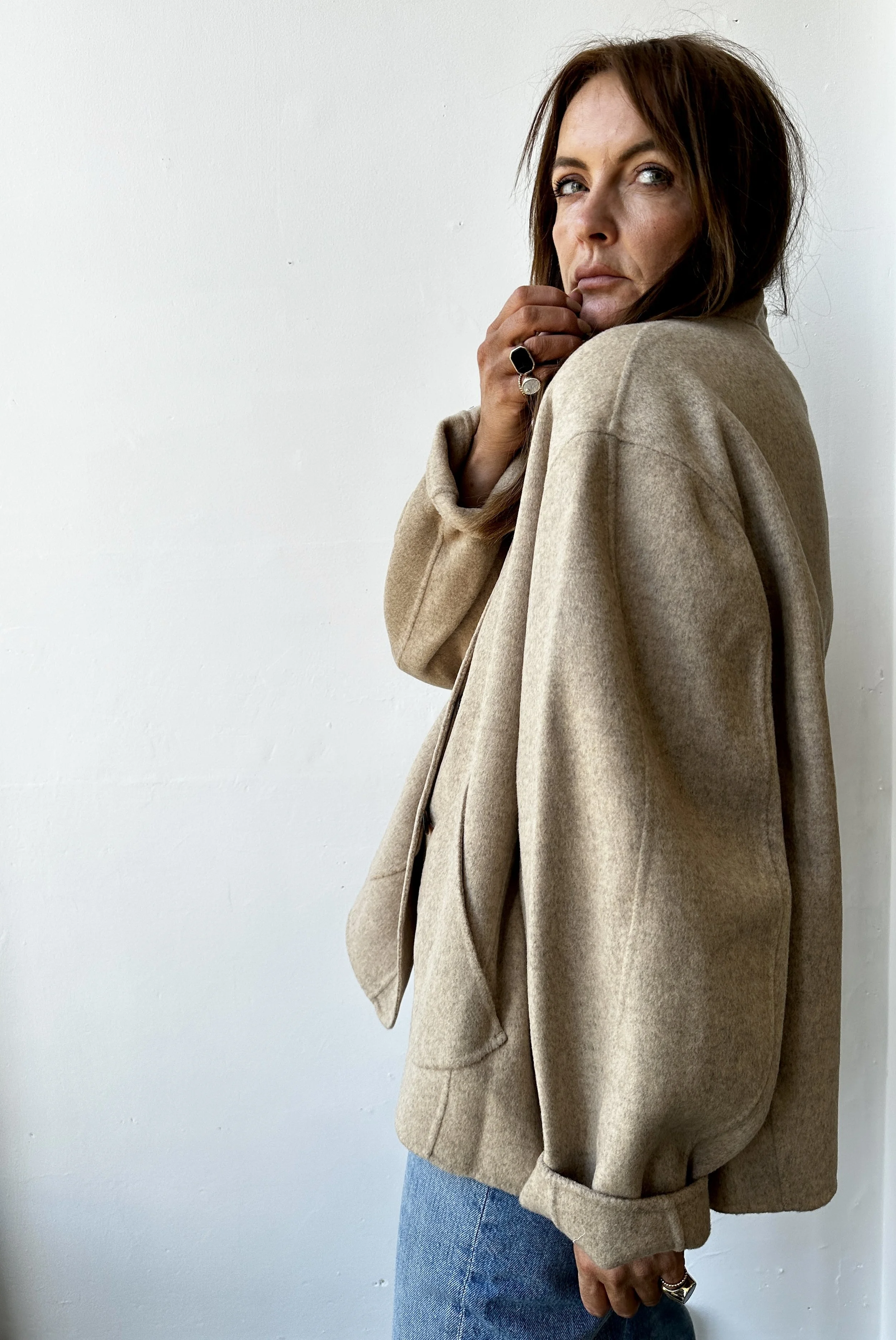 A woman with brown hair and blue eyes wearing a beige oversized blazer, blue jeans, and rings on her fingers, standing against a white wall, looking over her shoulder.