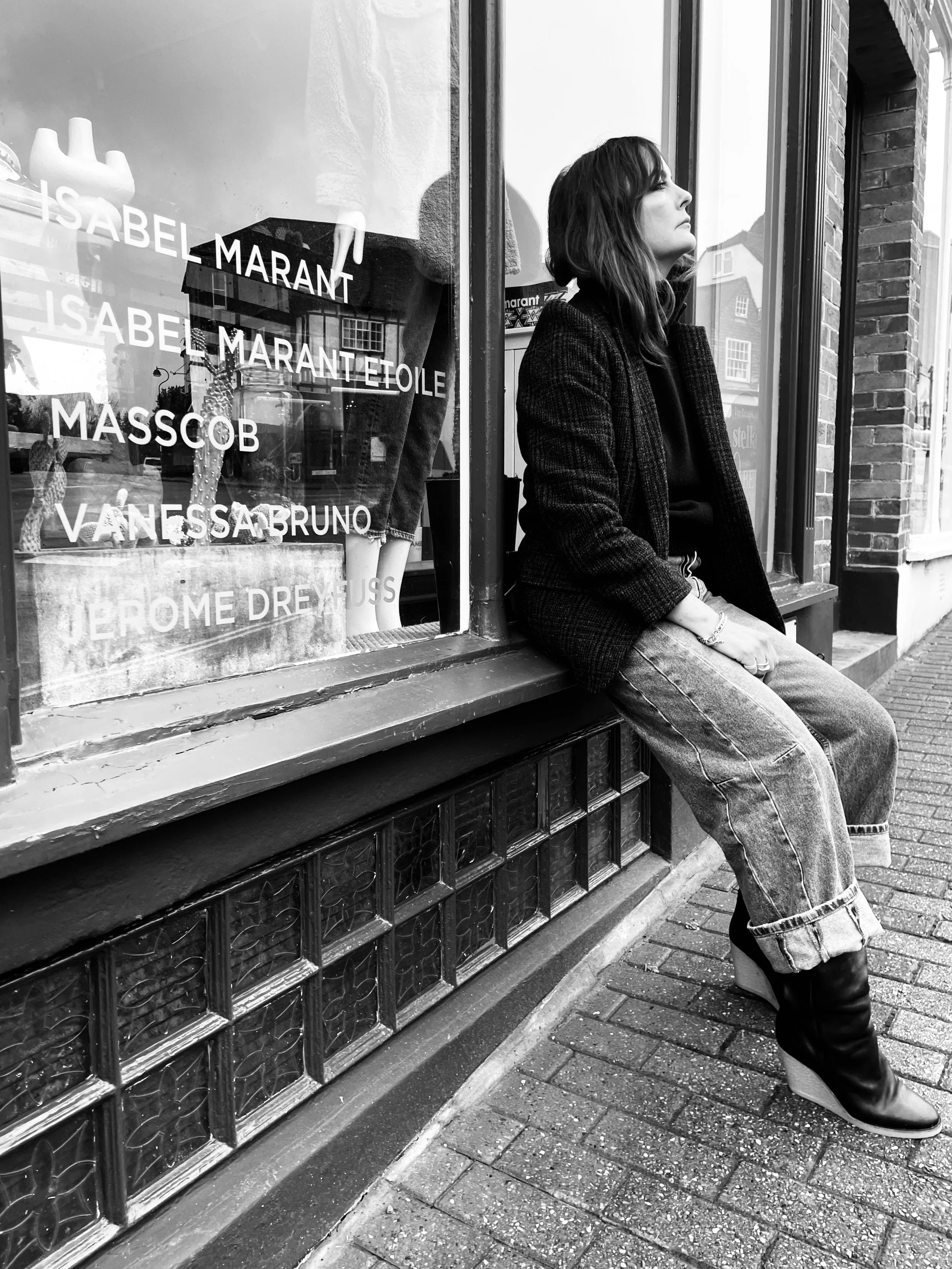 A woman sitting on a windowsill outside a storefront with clothing and designer names displayed in the window, wearing a blazer, loose jeans, and high-heeled boots in black and white.