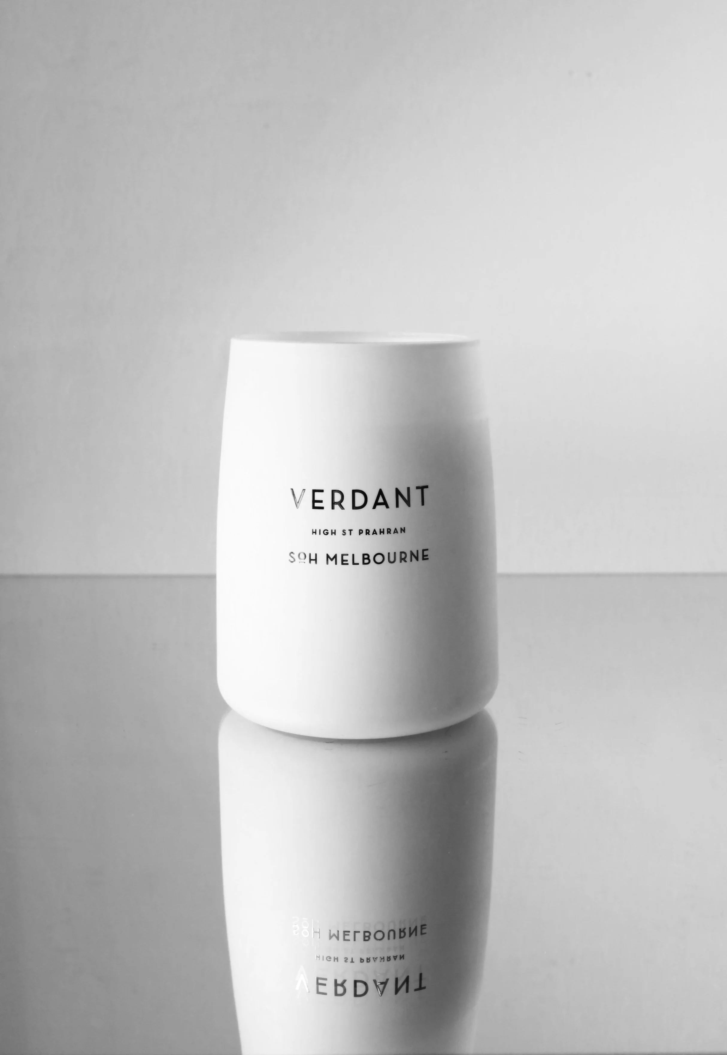 A white ceramic cup with black text that reads 'Verdant, High St Prahran, South Melbourne' is placed on a reflective surface, creating a mirror image of the cup and text.