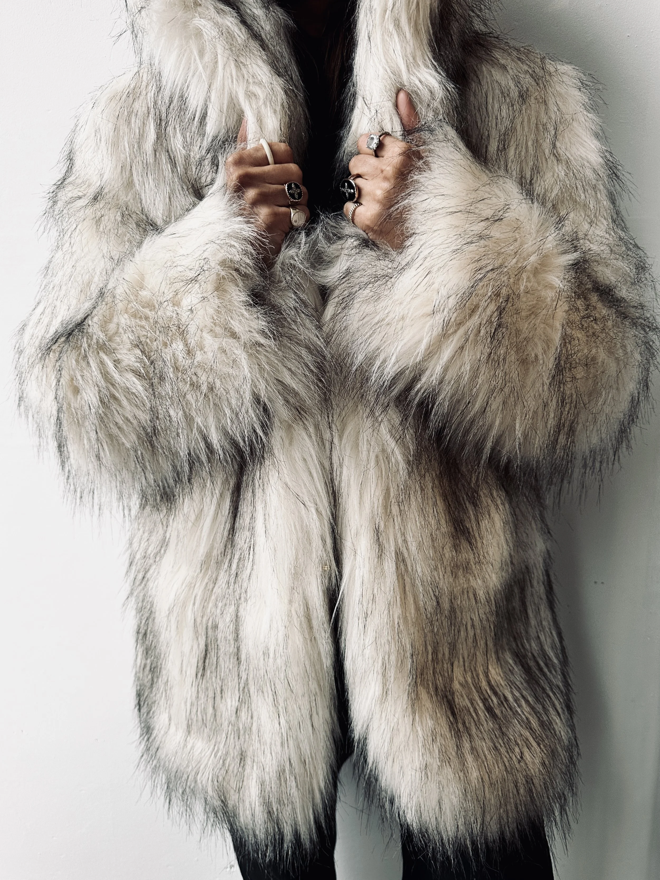 Person wearing a fur coat, showing rings on their fingers, against a plain background.
