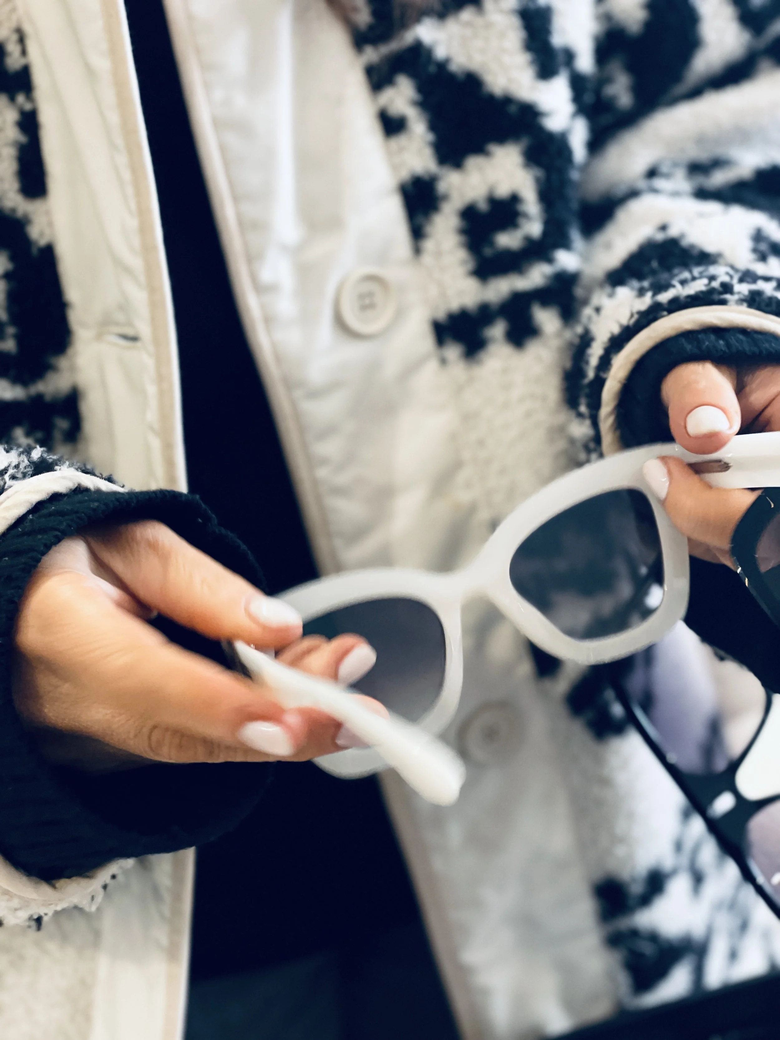 Person holding a white pair of sunglasses with black lenses, wearing a beige and black patterned sweater and a cream-colored vest.