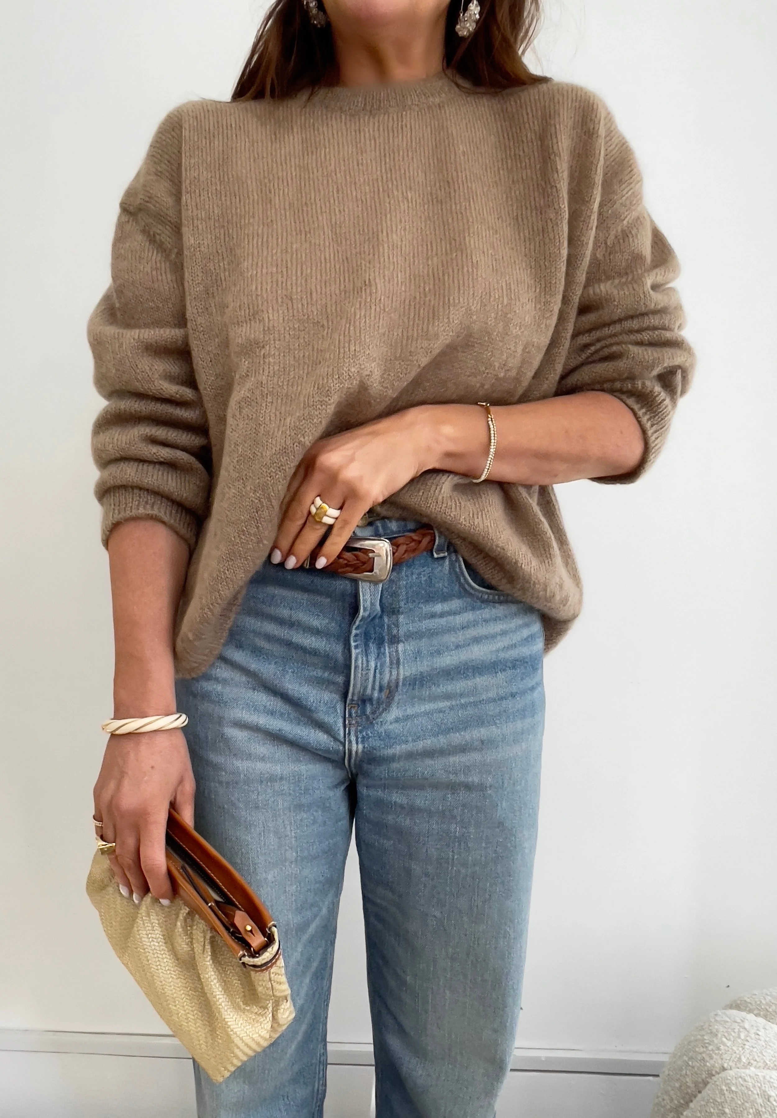 A woman wearing a beige oversized sweater, light blue jeans, and accessorized with gold and silver jewelry, holding a cream-colored clutch purse.