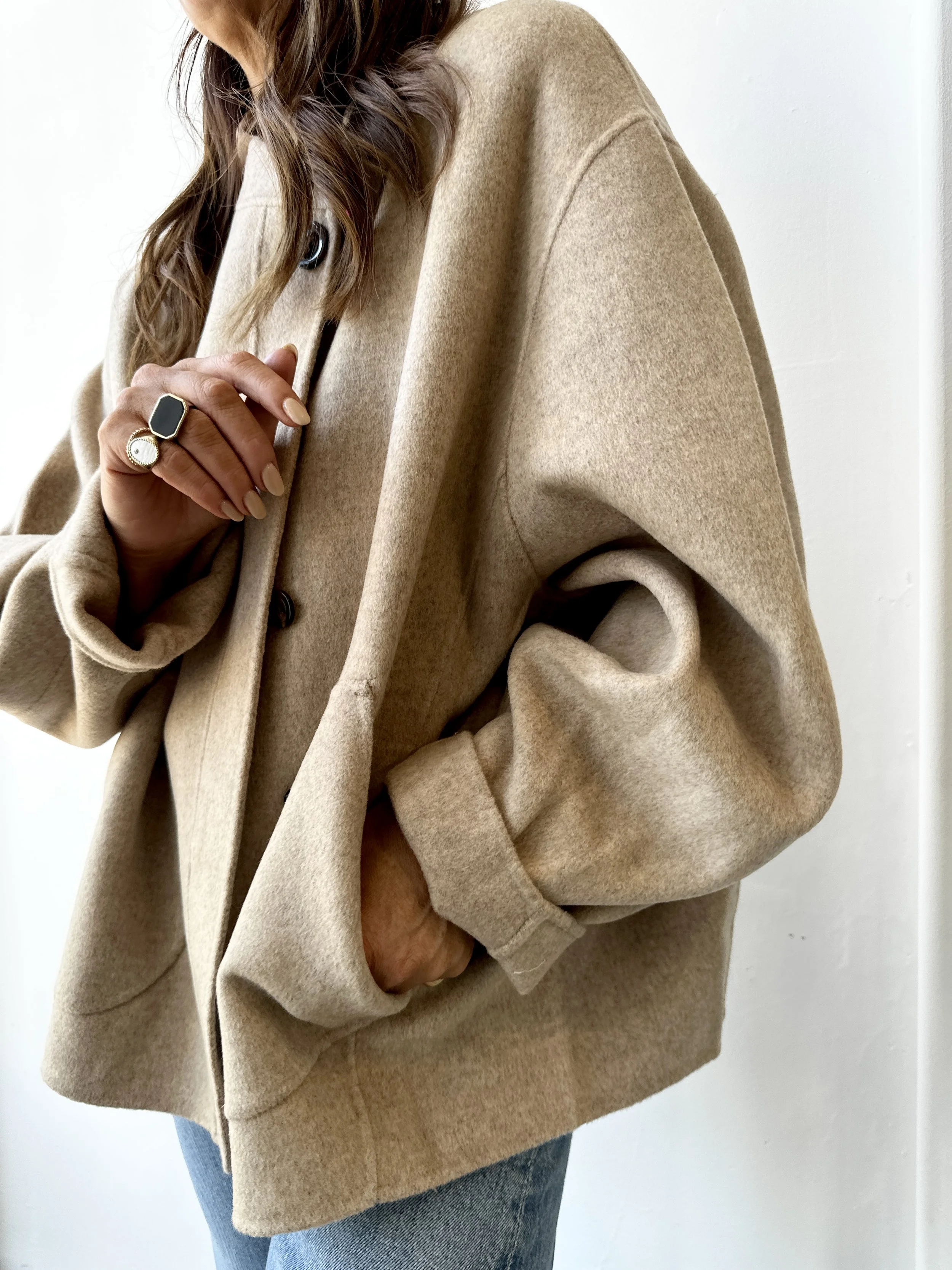 Person wearing a beige oversized coat with their hand put in the pocket, showing rings on their fingers and a partial view of blue jeans.