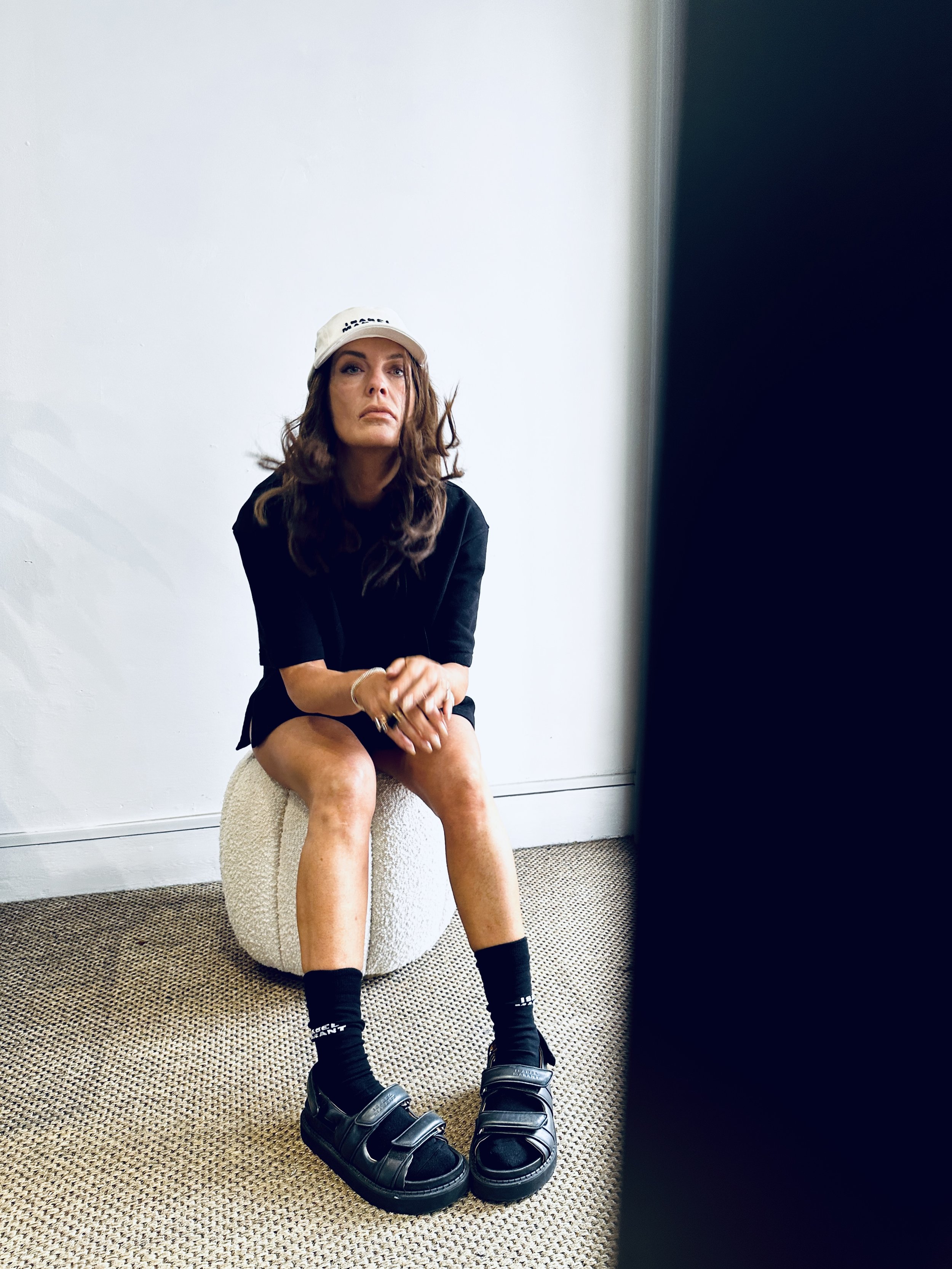 A woman with long brown hair sitting on a round textured stool in a corner of a room. She is wearing a white cap, black T-shirt, black socks, and chunky black sandals. The background is plain white with a dark object partially obscuring the right sid