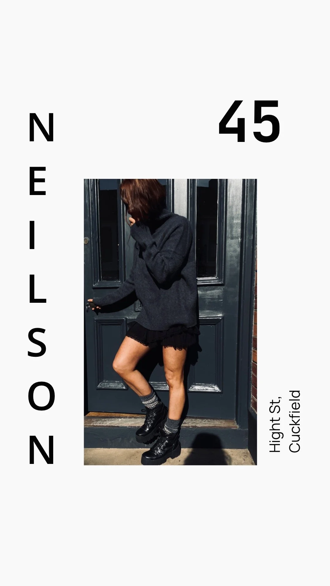 A person with red hair wearing a dark sweater, black shorts, gray socks, and black platform boots, standing in front of a blue door.