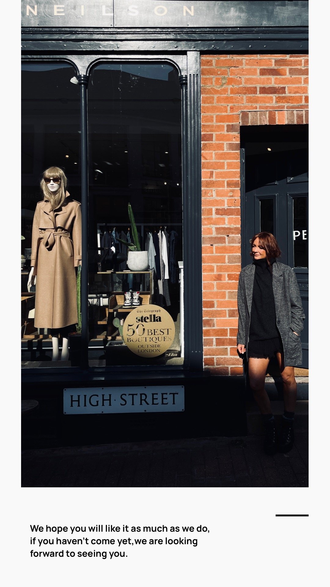 Image of a woman standing outside a boutique store with a mannequin in the window dressed in a tan coat and sunglasses. The store has a brick facade, black window frames, and a sign that reads 'HIGH STREET.' A round sign in the shop window advertises