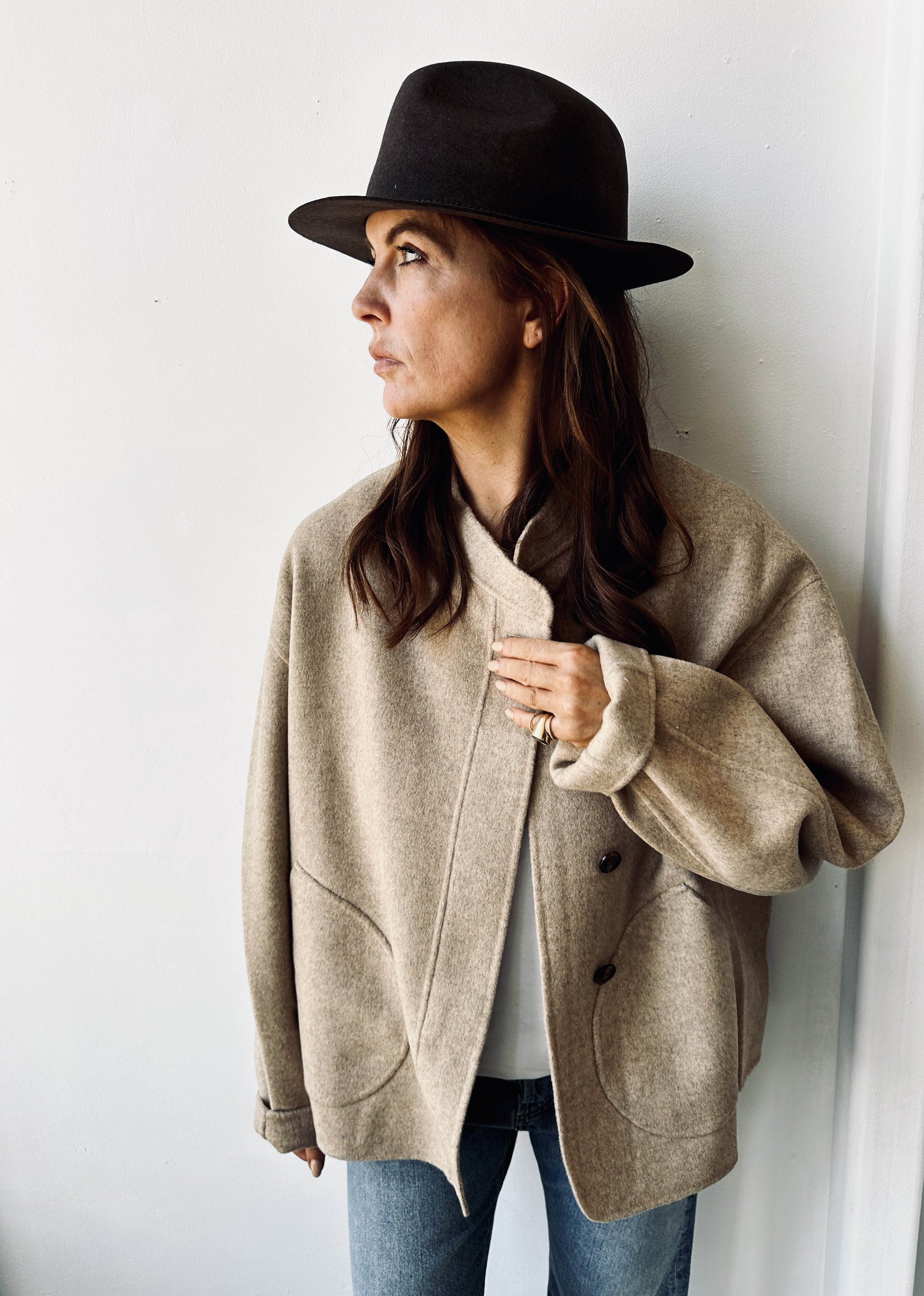 A woman wearing a beige coat and a black wide-brimmed hat is looking to the left, standing in front of a plain white wall.