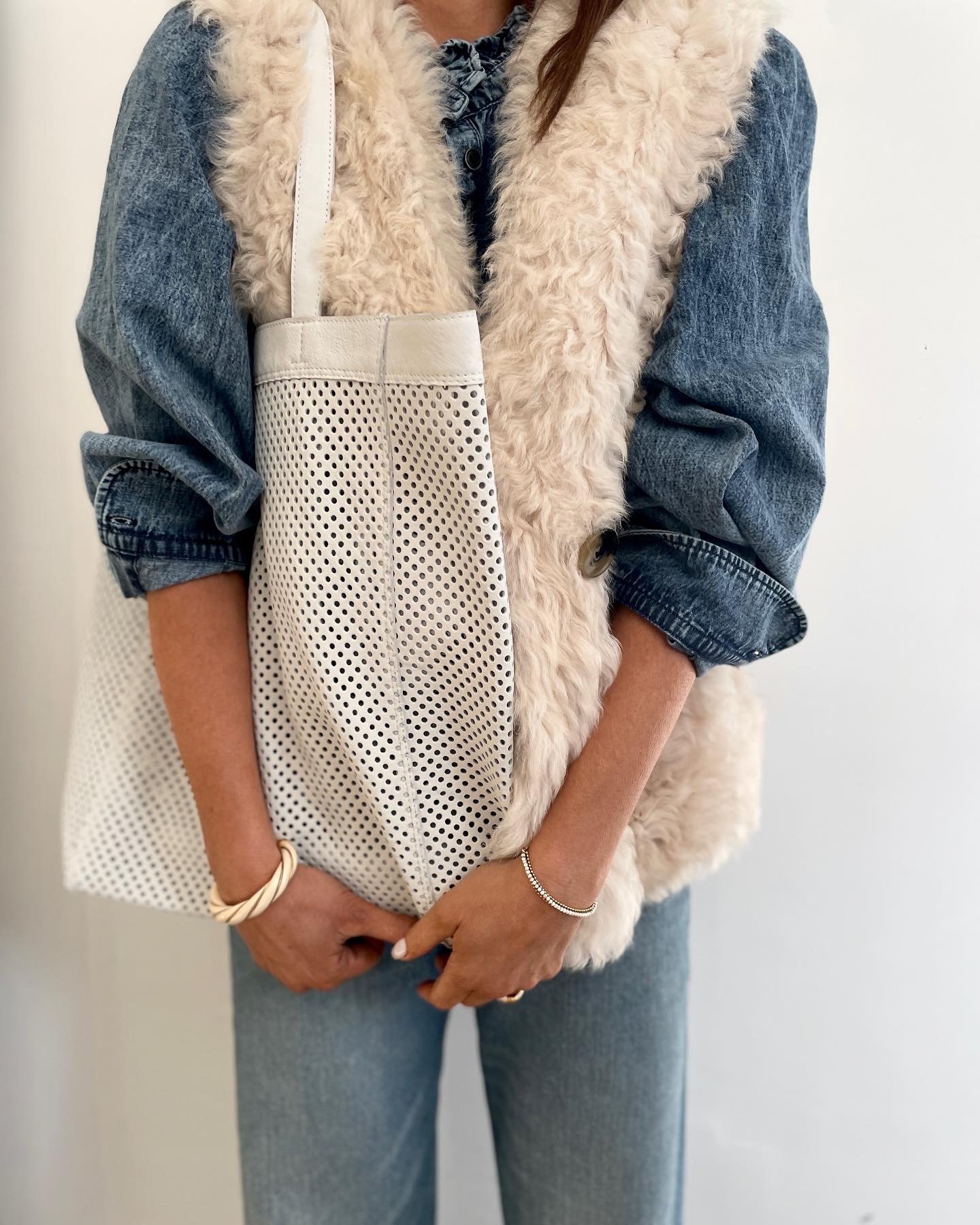 Person holding a perforated white tote bag, wearing a blue denim jacket with rolled-up sleeves, a beige faux fur vest, and blue jeans, with jewelry including a bracelet and a ring.