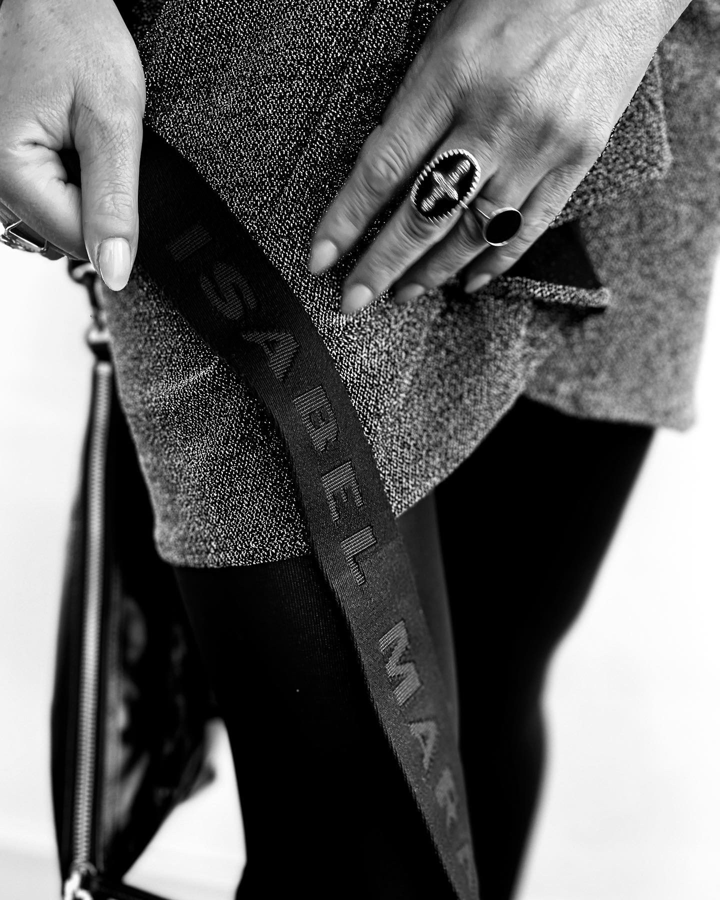 Close-up of a person's hand with rings holding the hem of a textured, heather gray garment with a black strap that has the word 'SEE BY' written on it, and black pants with a zipper detail.