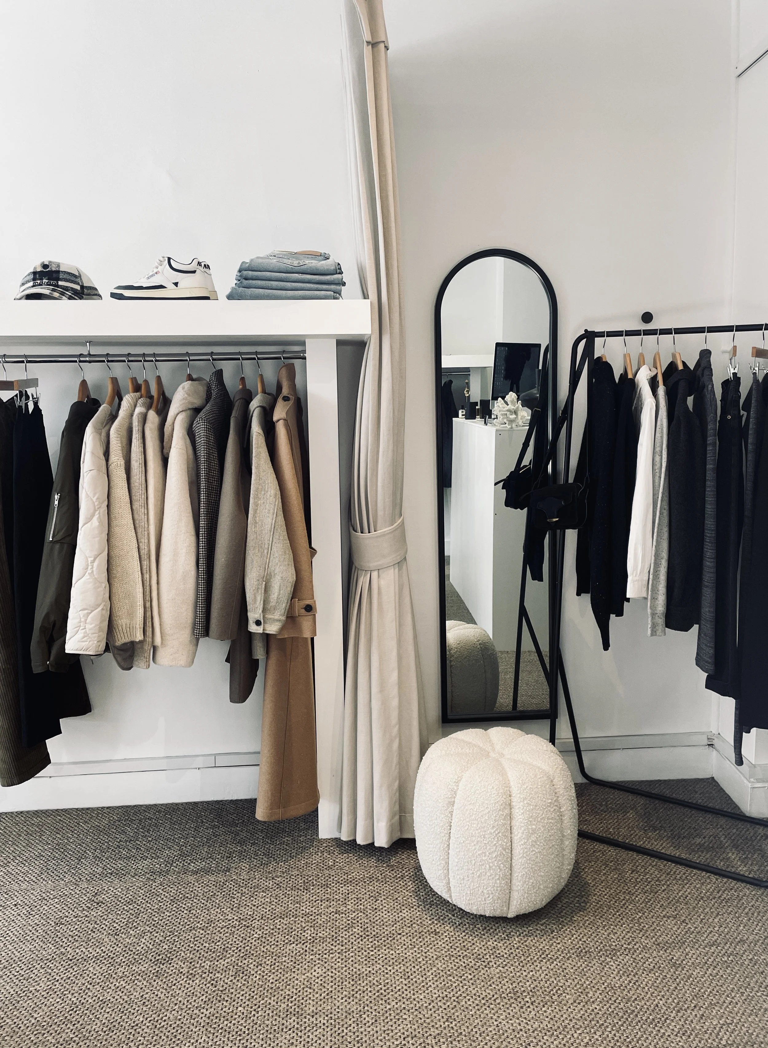 Clothing store display with jackets and shirts on racks, folded clothes on a shelf, a standing mirror, and a plush white ottoman.