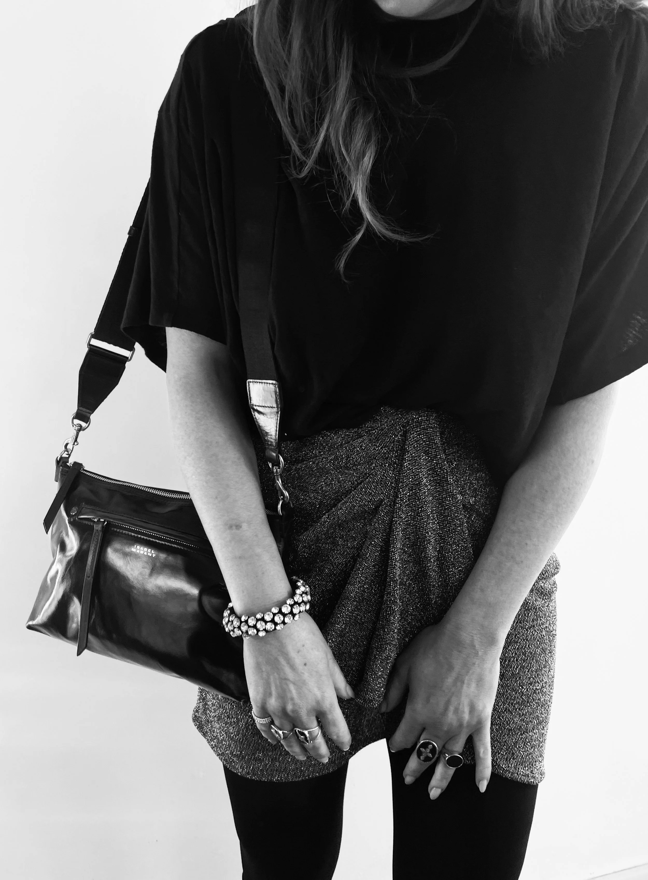 Black and white photo of a woman wearing a loose black top, a shimmery skirt, and a bracelet. She is holding a shiny black purse with a strap and has multiple rings on her fingers.