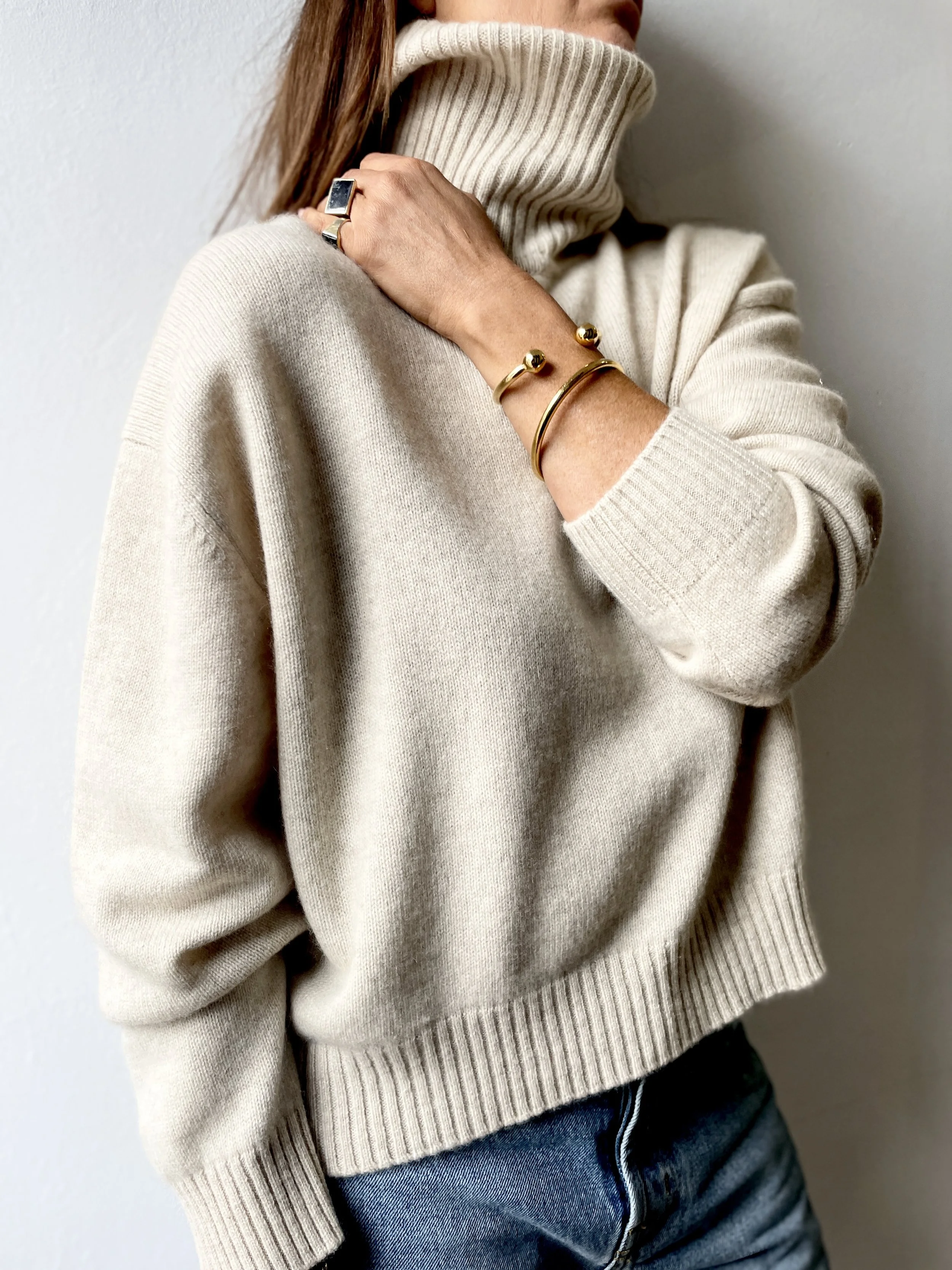 A woman in a cream-colored turtleneck sweater and jeans, showcasing jewelry including a silver ring and gold bracelets.