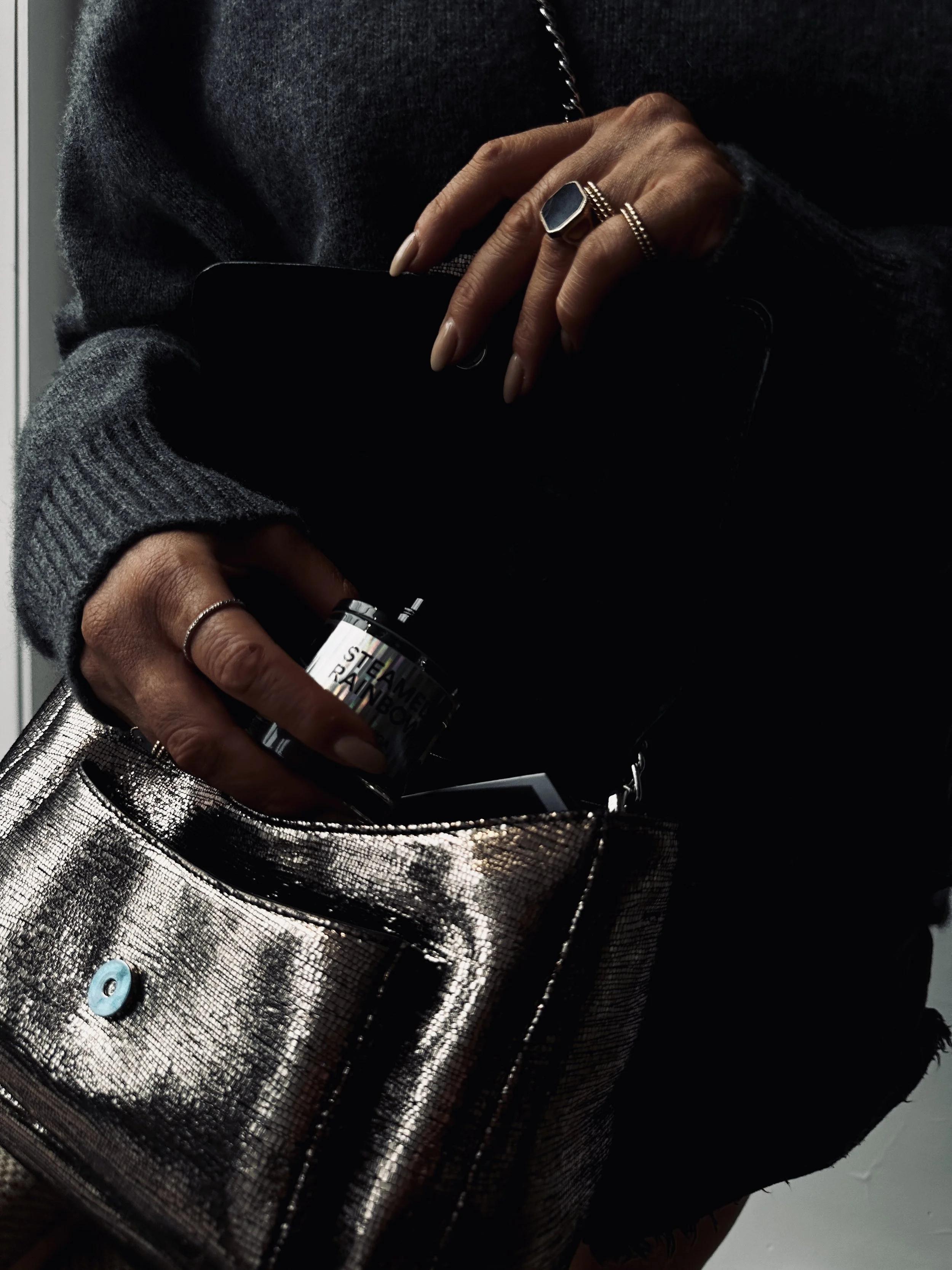A person reaching into a shiny metallic bag, holding a black container with the words 'Steamery Rainbow' printed on it, while wearing multiple rings and a gray sweater.