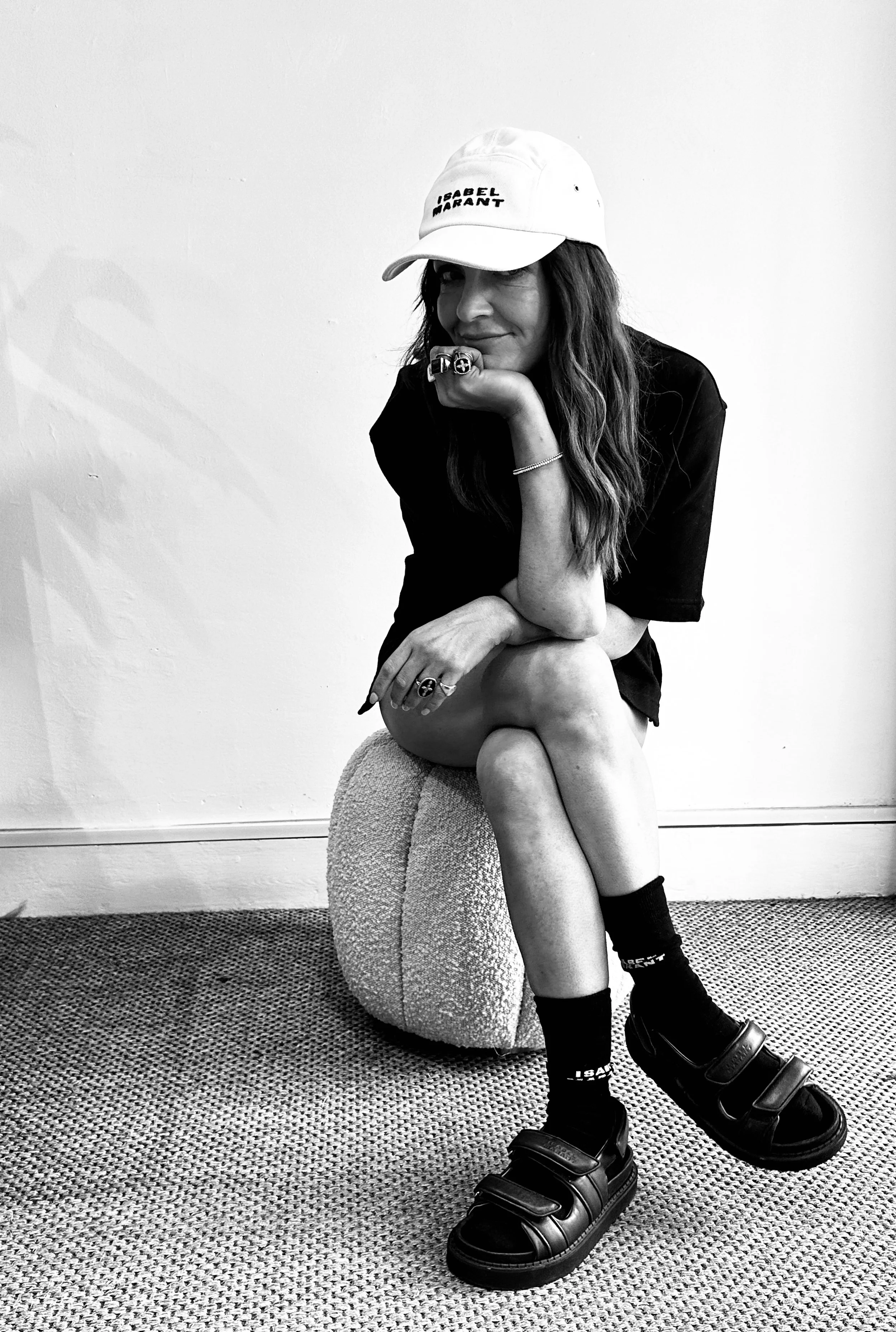 A woman with long hair wearing a white cap, black t-shirt, socks, and sandals sitting on a textured ottoman against a plain wall. She is resting her chin on her hand and winking at the camera.