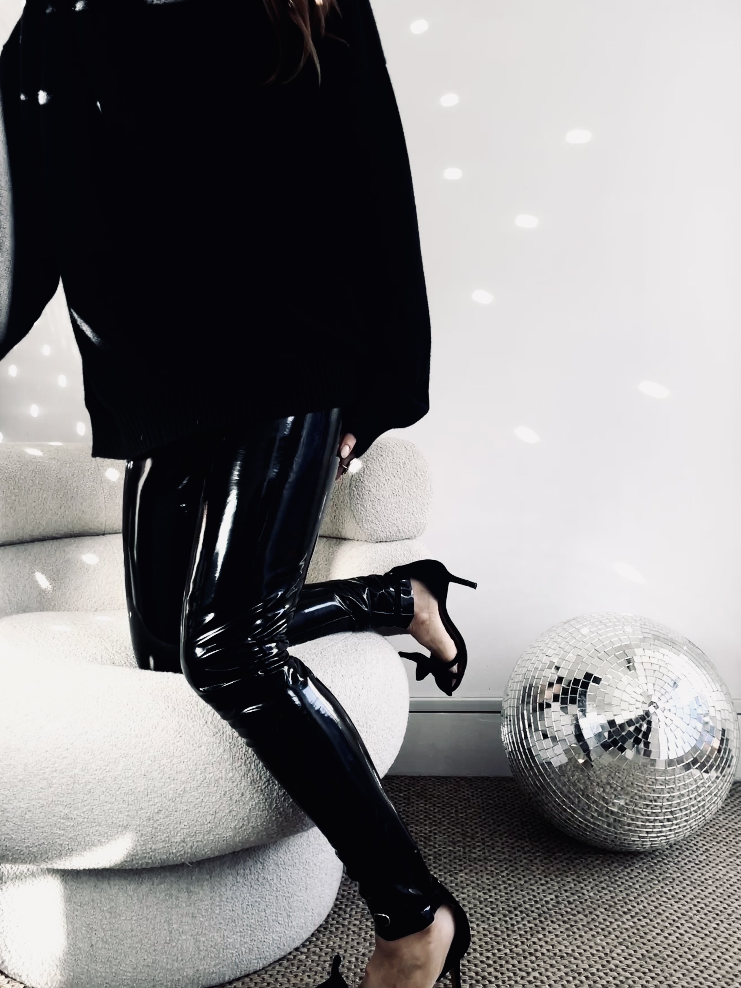 A person in black shiny latex pants, black top, and black high heels is kneeling on a textured beige sofa with one knee on the cushion and the other foot on the ground. A large disco ball is on the carpeted floor nearby, and there are small white lig