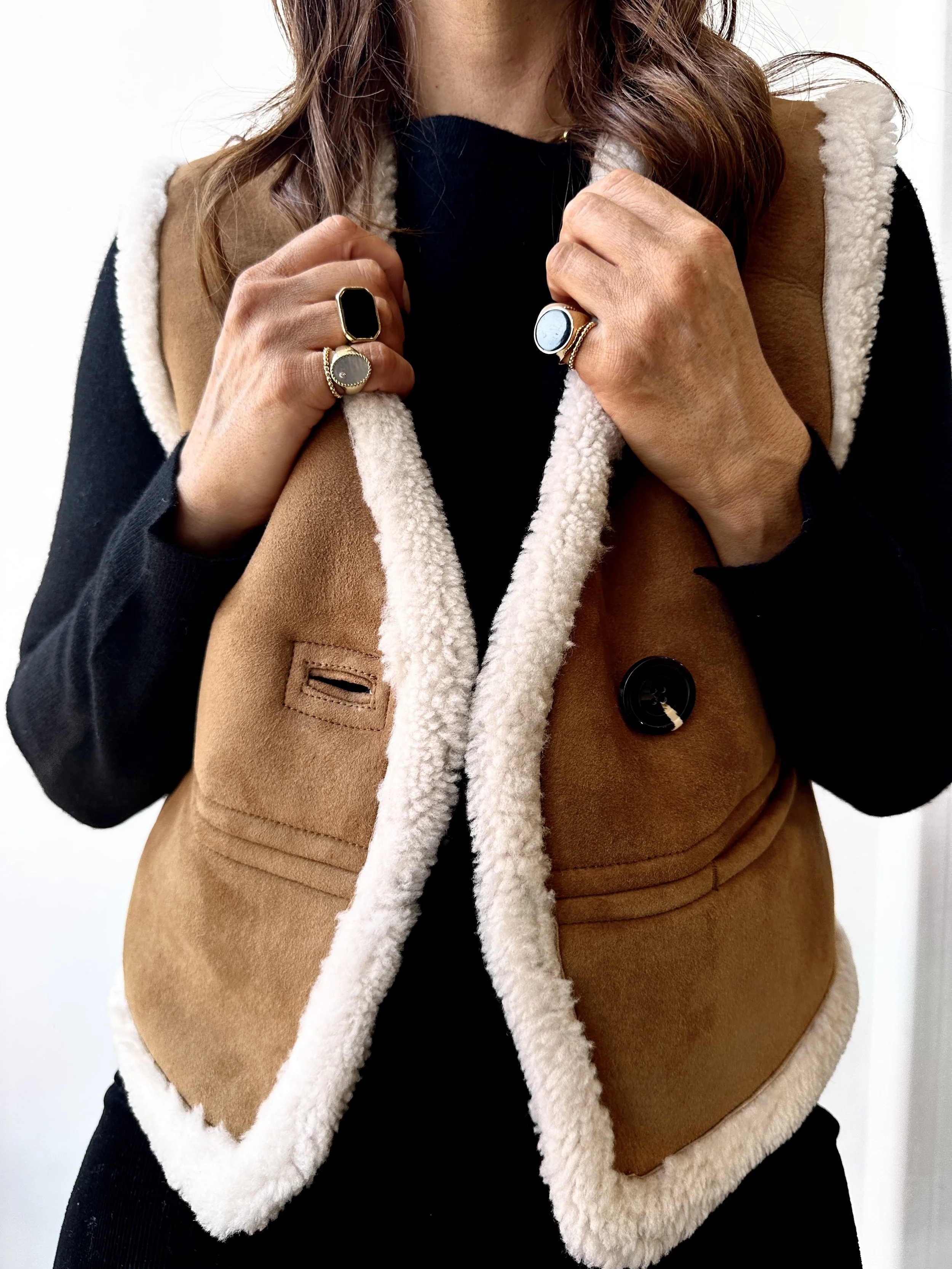 A woman wearing a black long-sleeve shirt and a tan fleece-lined vest, holding the collar of the vest with both hands. She is wearing multiple rings including a black stone ring, a silver ring with a circular face, and a gold ring with a black inset.