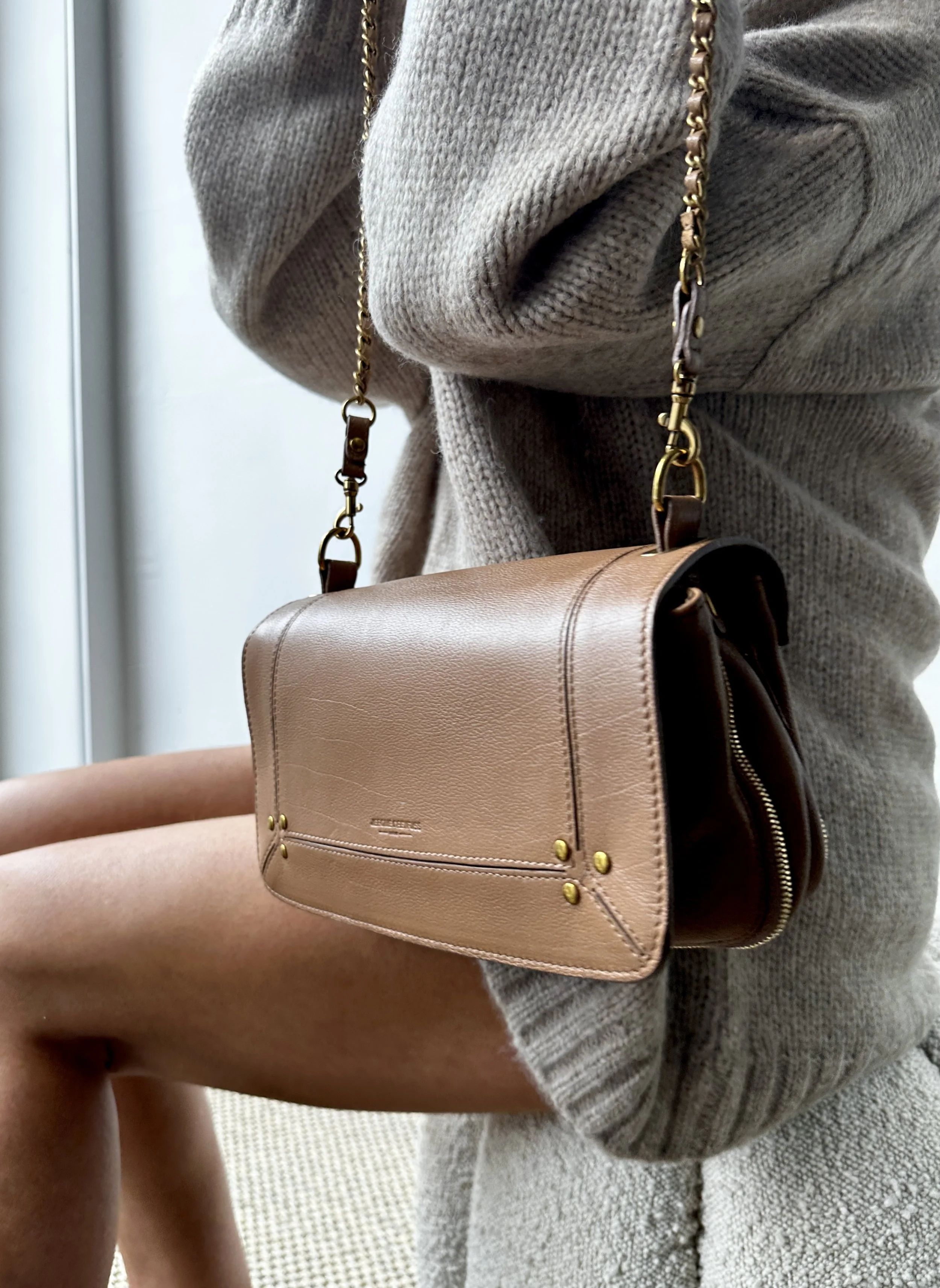 A beige leather handbag with gold accents, hanging from a gold chain strap, worn by a person in a gray sweater and textured skirt.