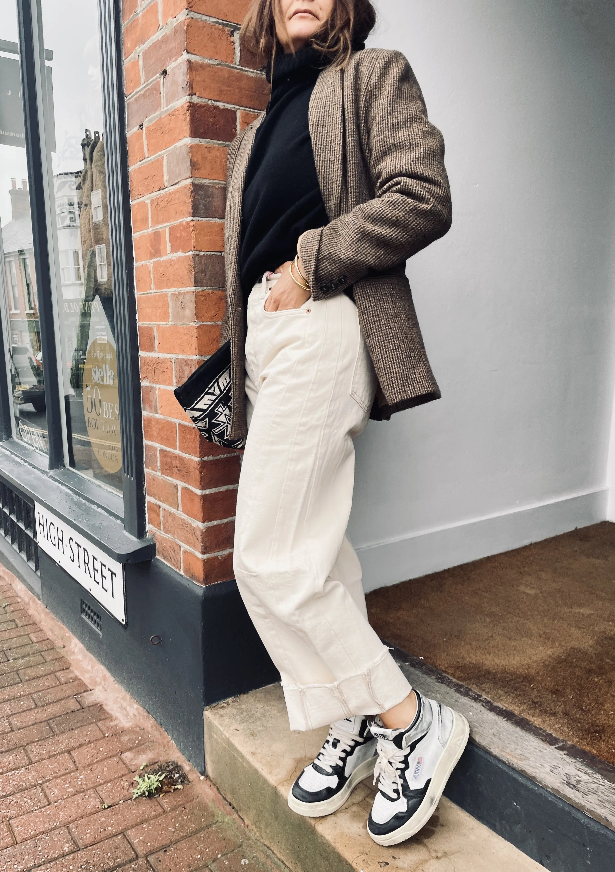 A woman with a plaid blazer, black turtleneck, and wide beige pants standing against a brick wall outside a building with a street sign that reads 'High Street'.
