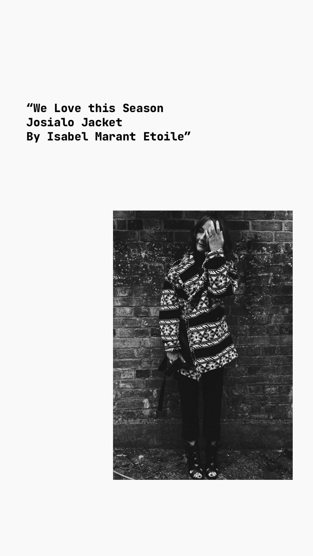 Black and white photo of a woman standing against a brick wall, wearing a patterned jacket, black pants, and open-toe shoes, with one hand touching her face.