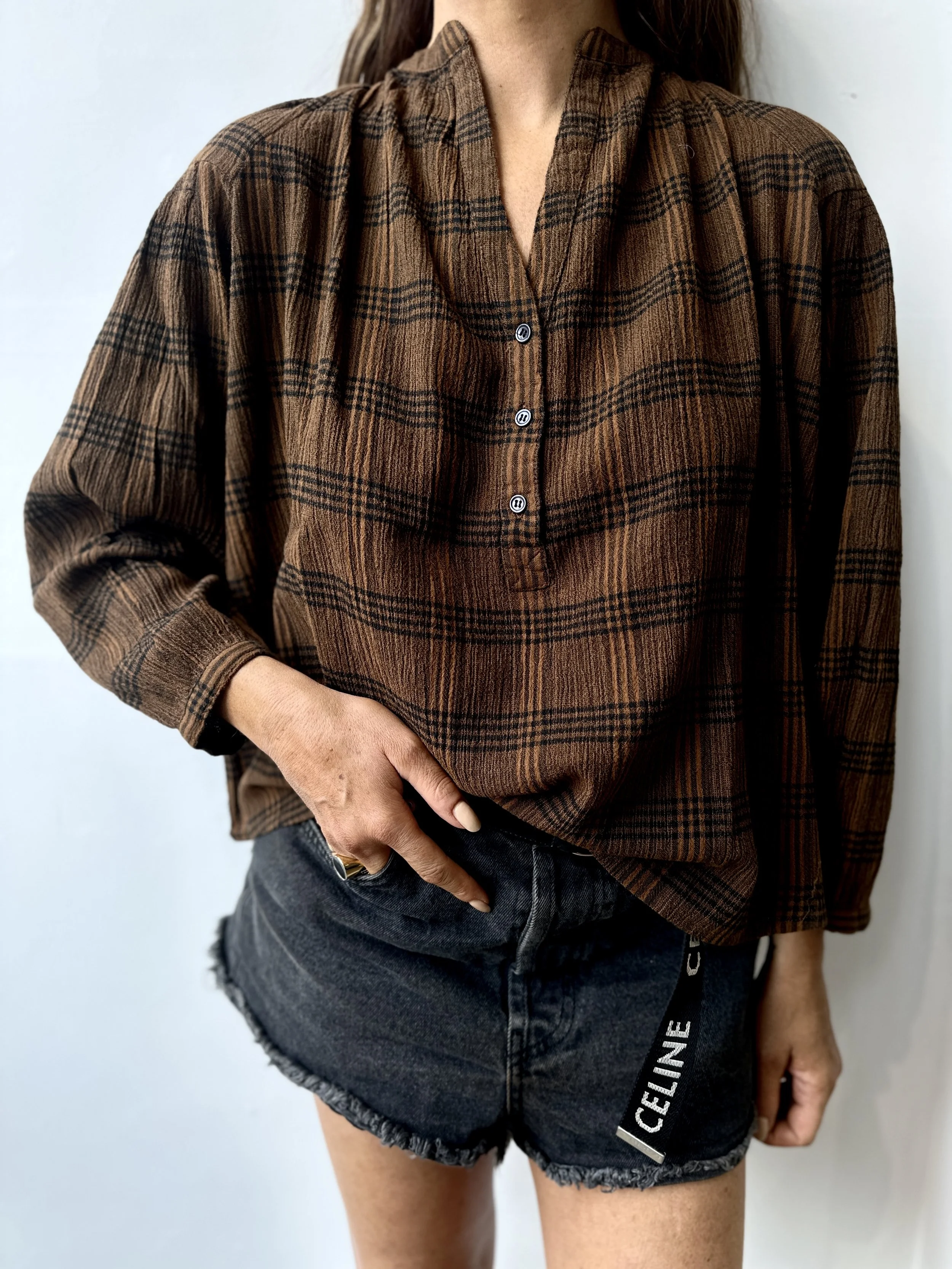 A woman wearing a brown plaid long-sleeve shirt and black denim shorts with a Celine label, standing against a plain background.