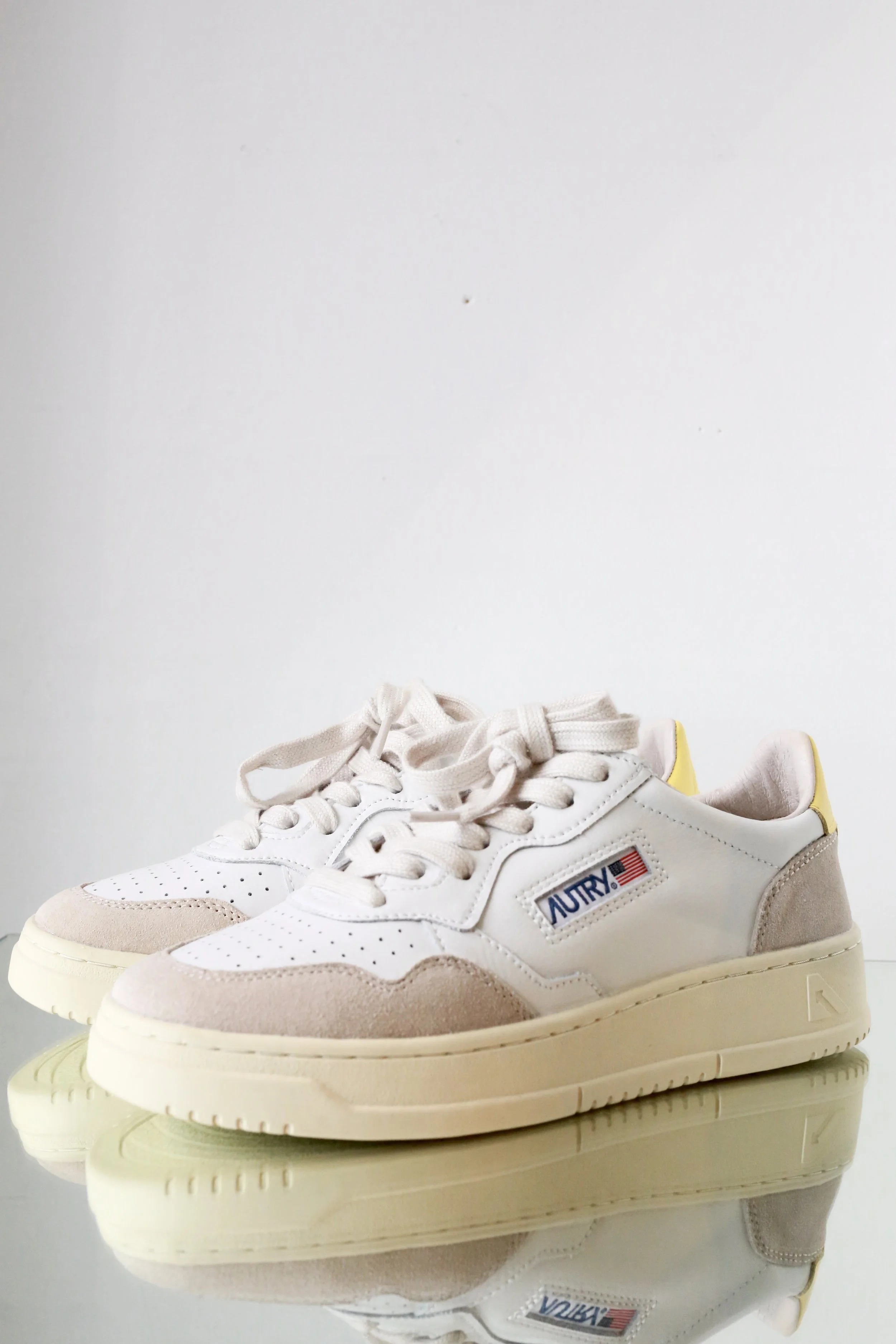 A pair of white and beige sneakers with a patch that reads 'AURY' and an American flag, placed on a reflective surface against a plain white background.