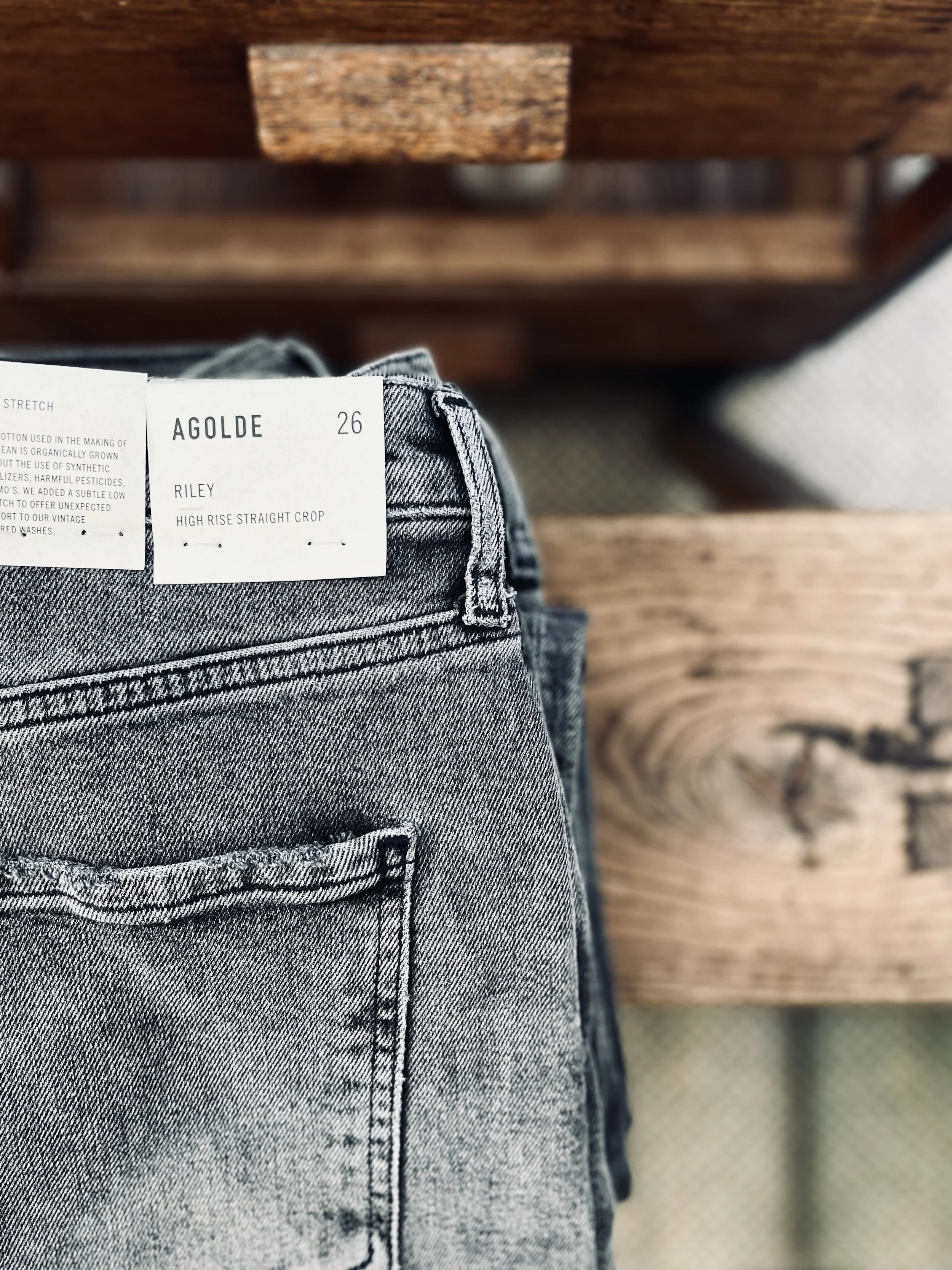 Gray high-rise straight crop jeans with a white label reading 'AGolde,' 'Riley,' size 26, on a person standing in front of a wooden surface.