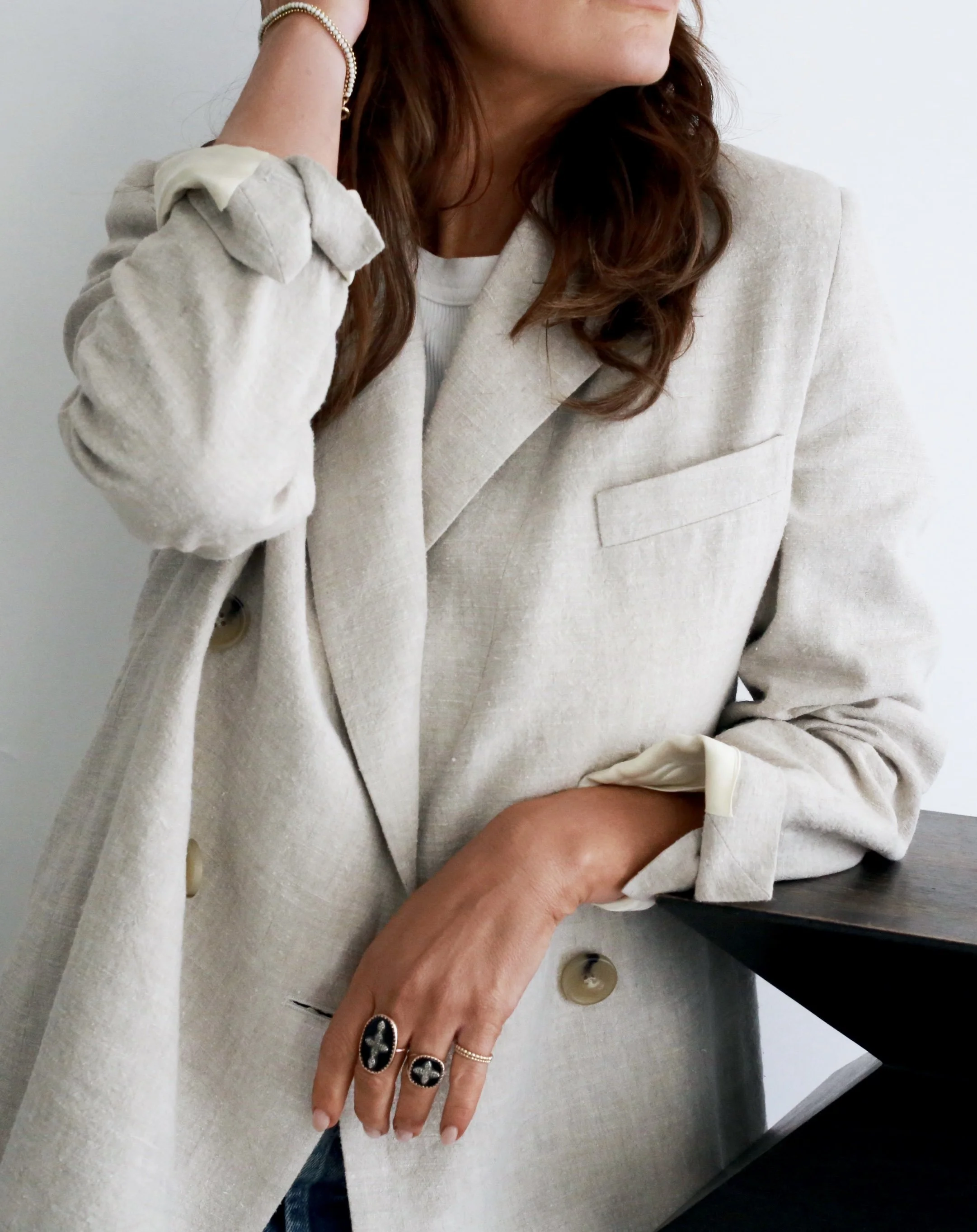 Woman wearing a light beige blazer with rolled-up sleeves, accessorized with rings and a bracelet, standing with her hand resting on a dark surface, against a plain white background.