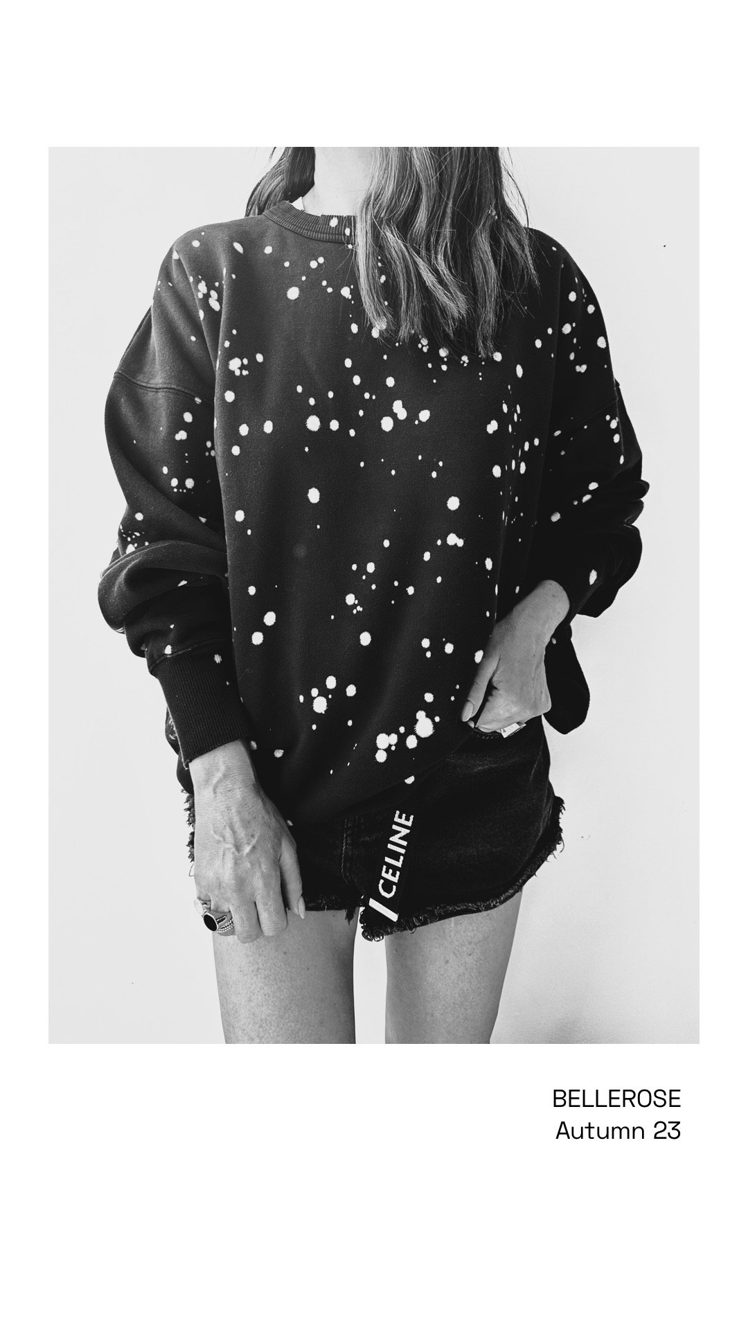 A person wearing a black sweatshirt with a white splatter pattern, black denim shorts with the word "CELINE" on the side, and holding a ring, facing a plain background. The text reads "BELLROSE Autumn 23."