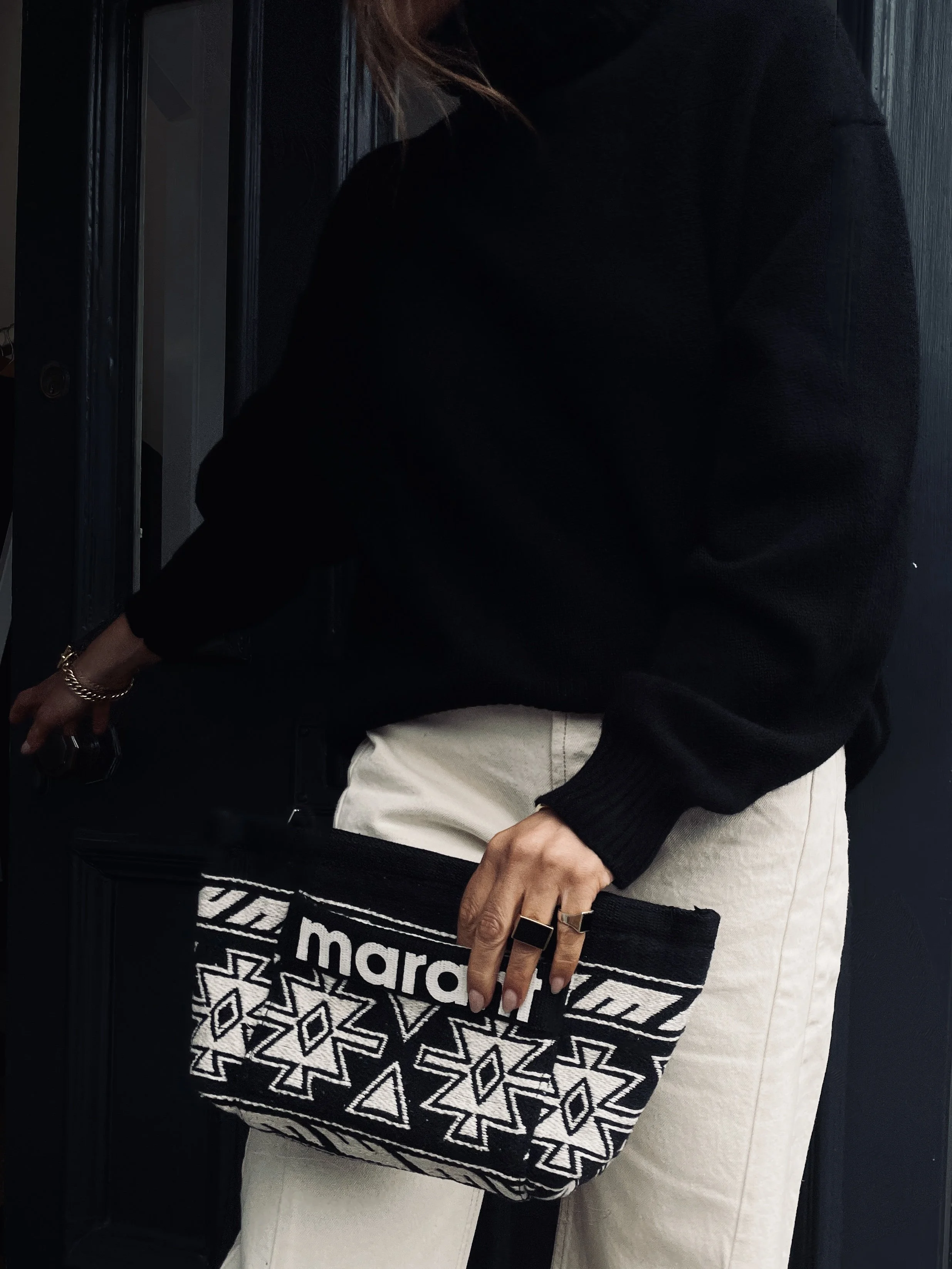 Person holding a black and white patterned Marais clutch purse, wearing white pants and a black sweater, with a gold bracelet and ring.