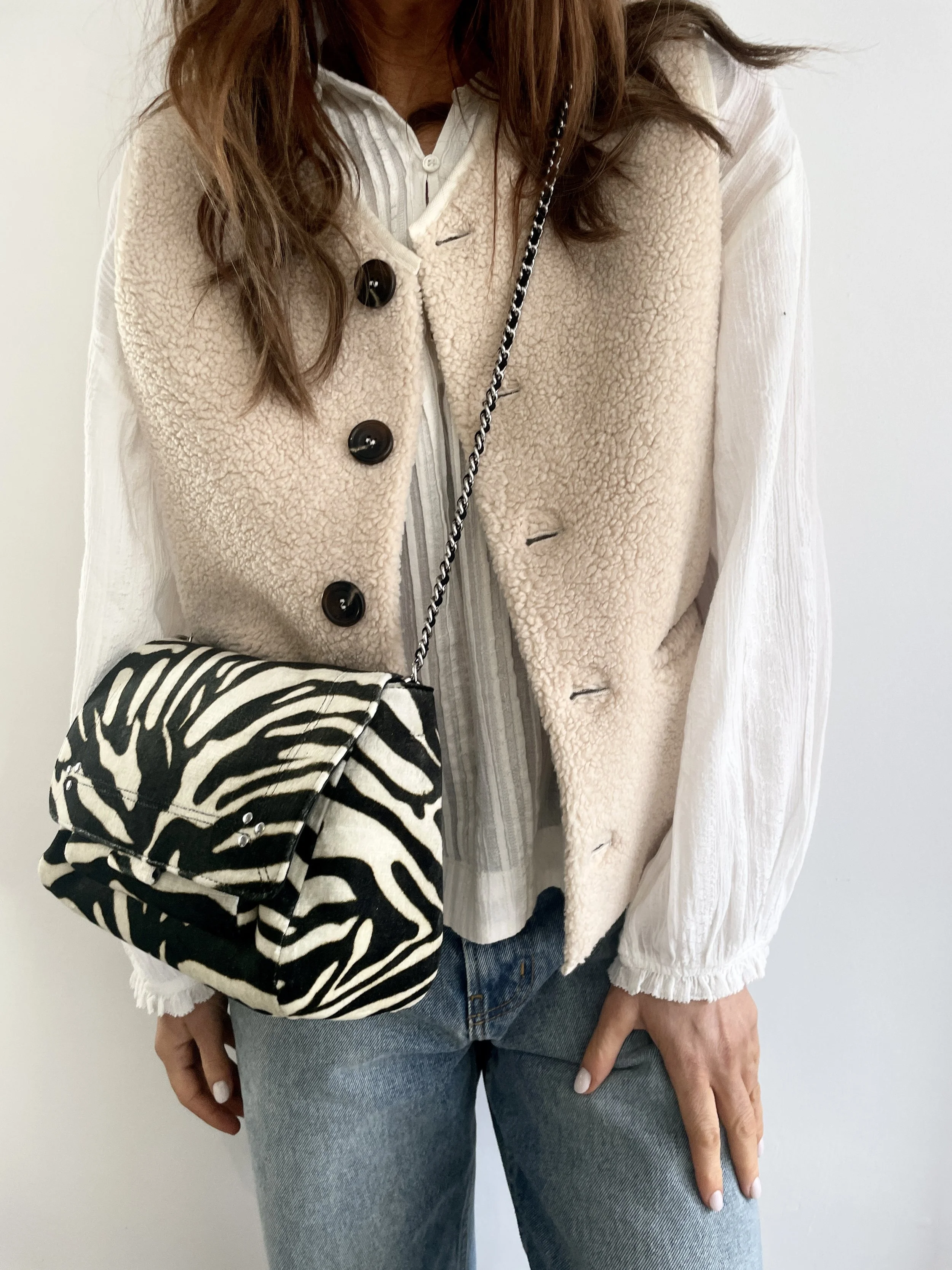 Person wearing a beige, textured vest over a white, long-sleeve blouse, carrying a black and white zebra print handbag with a chain strap, and wearing blue jeans.
