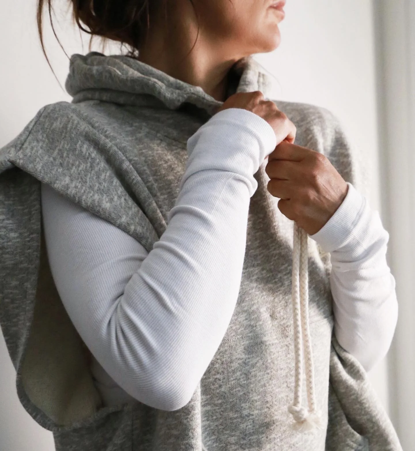 A woman wearing a gray hoodie and a white long sleeve shirt adjusting the collar of her hoodie.
