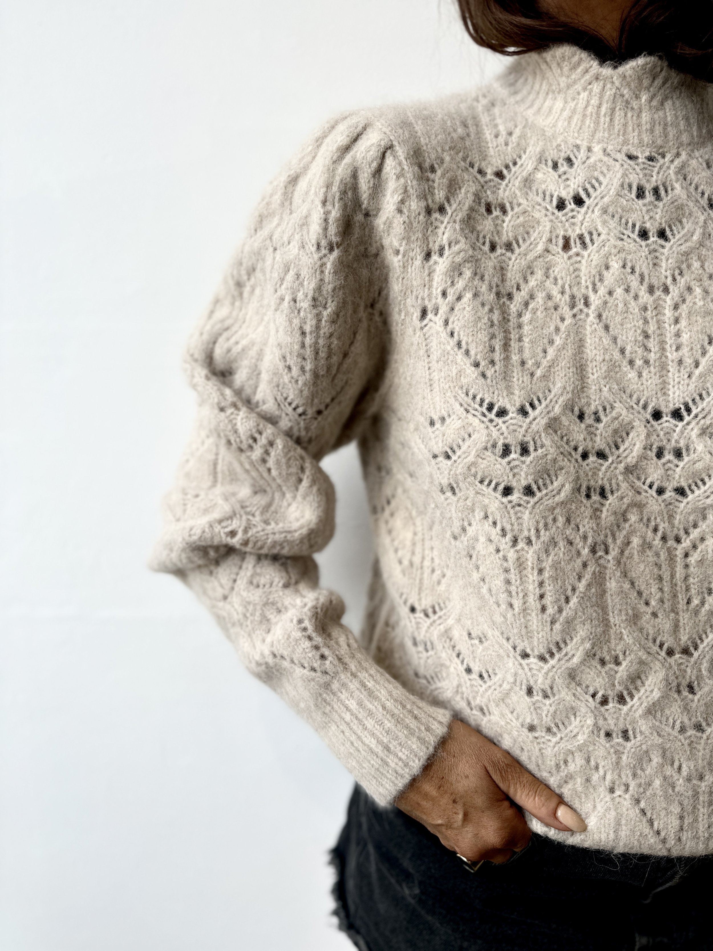 Woman wearing a beige knitted sweater with an intricate open lace pattern and a high neckline, standing against a plain white background.