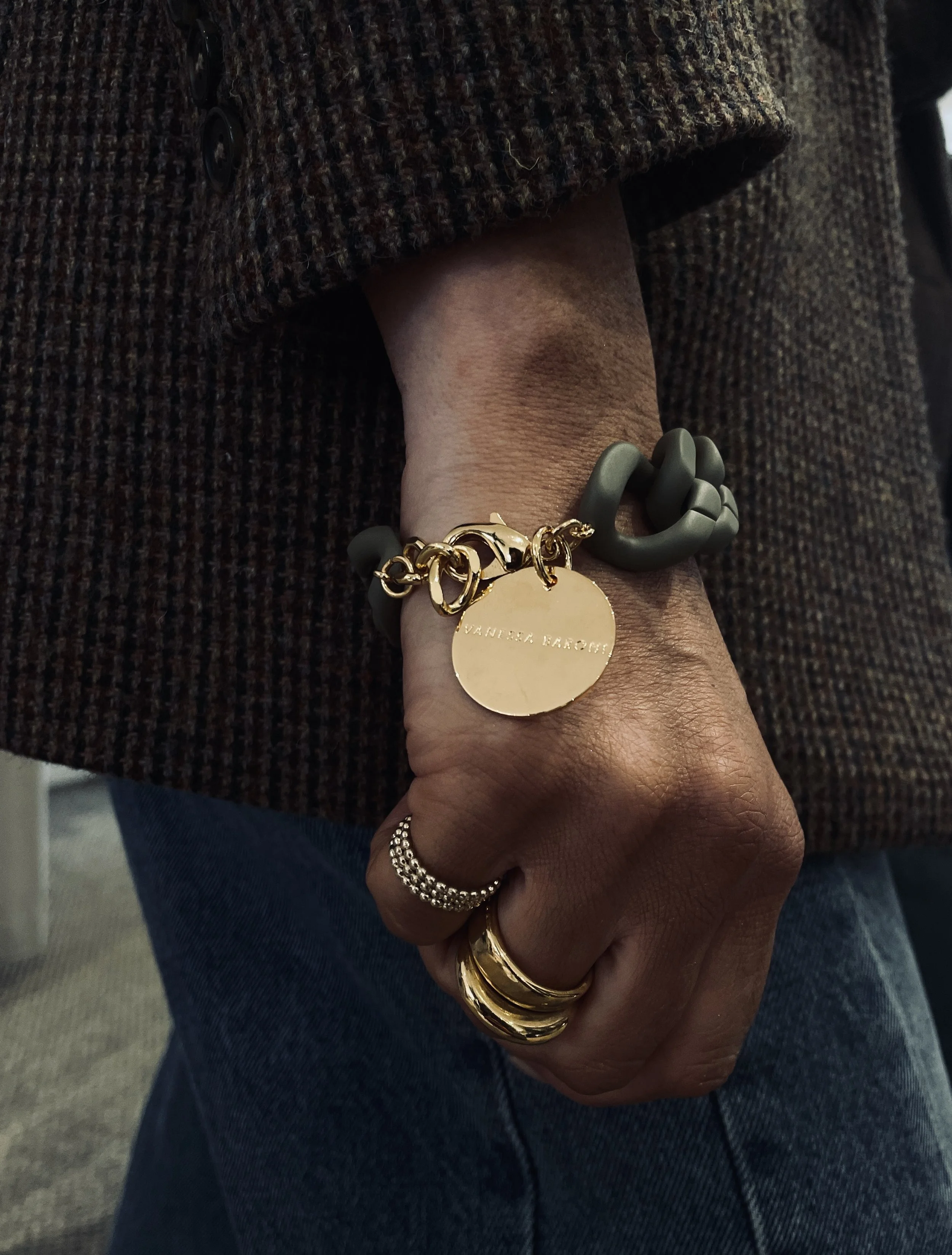Close-up of a person's hand wearing multiple rings and bracelets, including a gold chain bracelet with a round gold pendant engraved with 'VANESSA BRON' and a gray chunky knot bracelet, wearing a brown plaid coat and blue jeans.