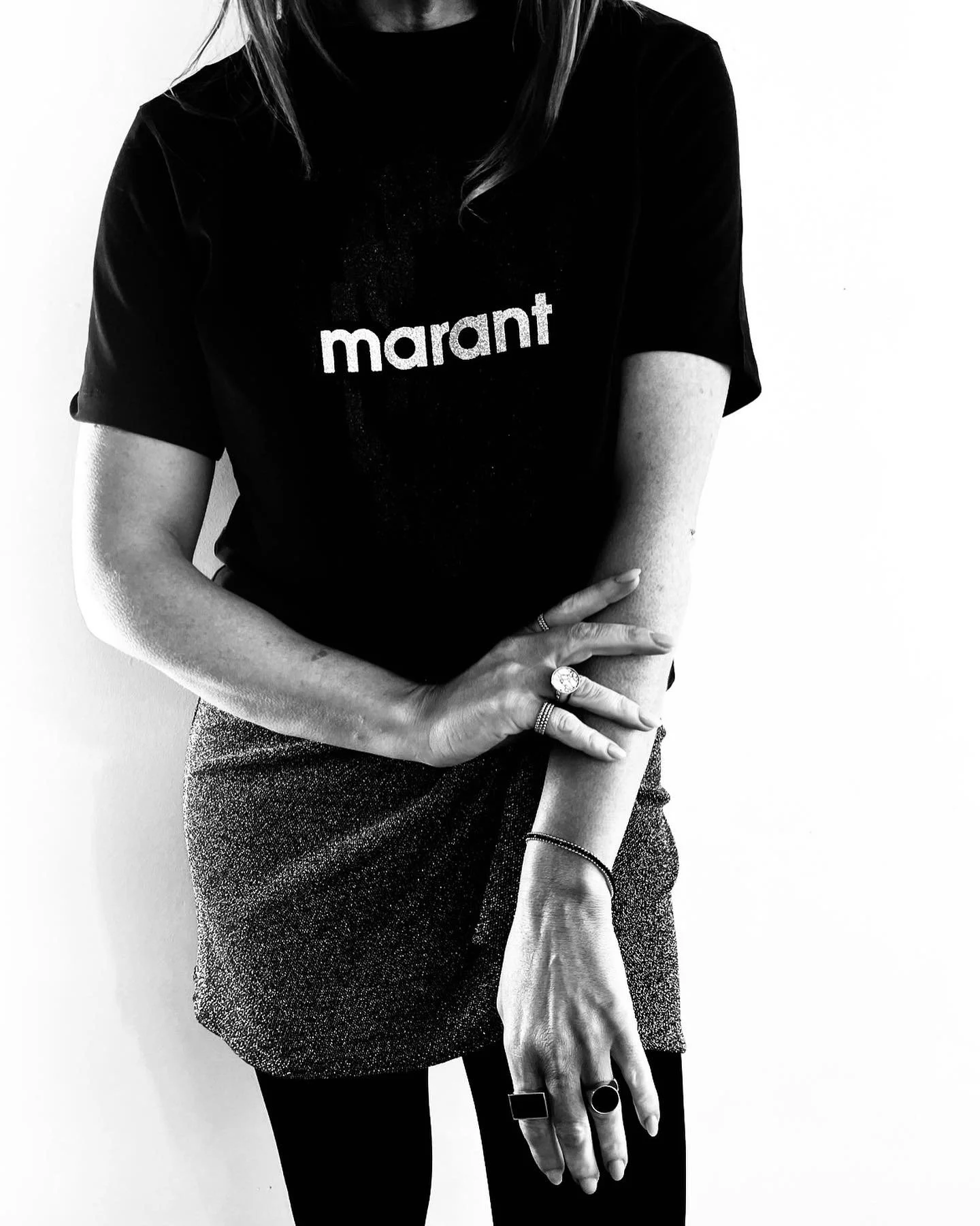 Black and white photo of a woman wearing a black shirt with the word 'marant' printed on it, and a textured skirt, standing against a plain white background. Her arms are crossed, and she is wearing multiple jewelry rings and bracelets.
