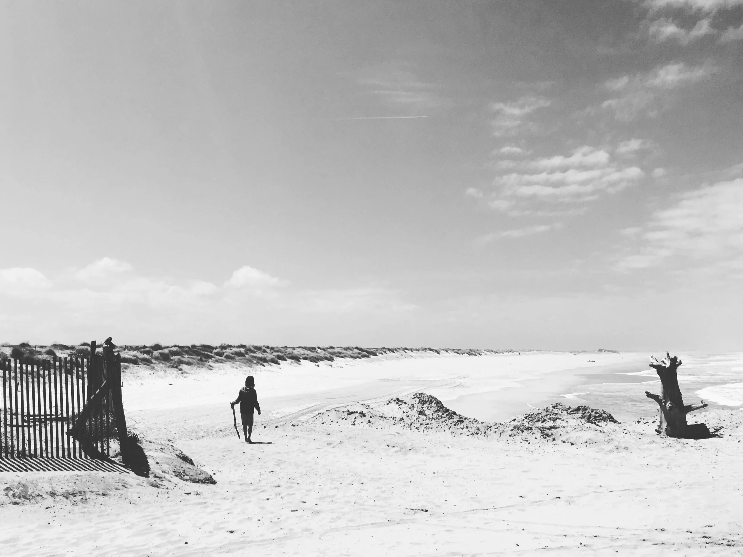 A black and white photo of a person walking along a sandy beach with a walking stick, near a fence and a piece of driftwood, with a vast sky and distant shoreline.