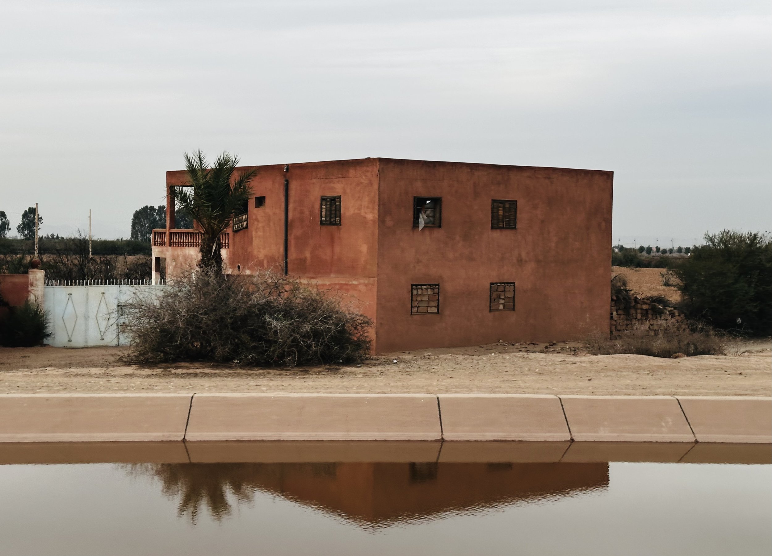A two-story, earth-toned house with small, barred windows, a palm tree nearby, and dry desert surroundings reflected in a water feature in the foreground.