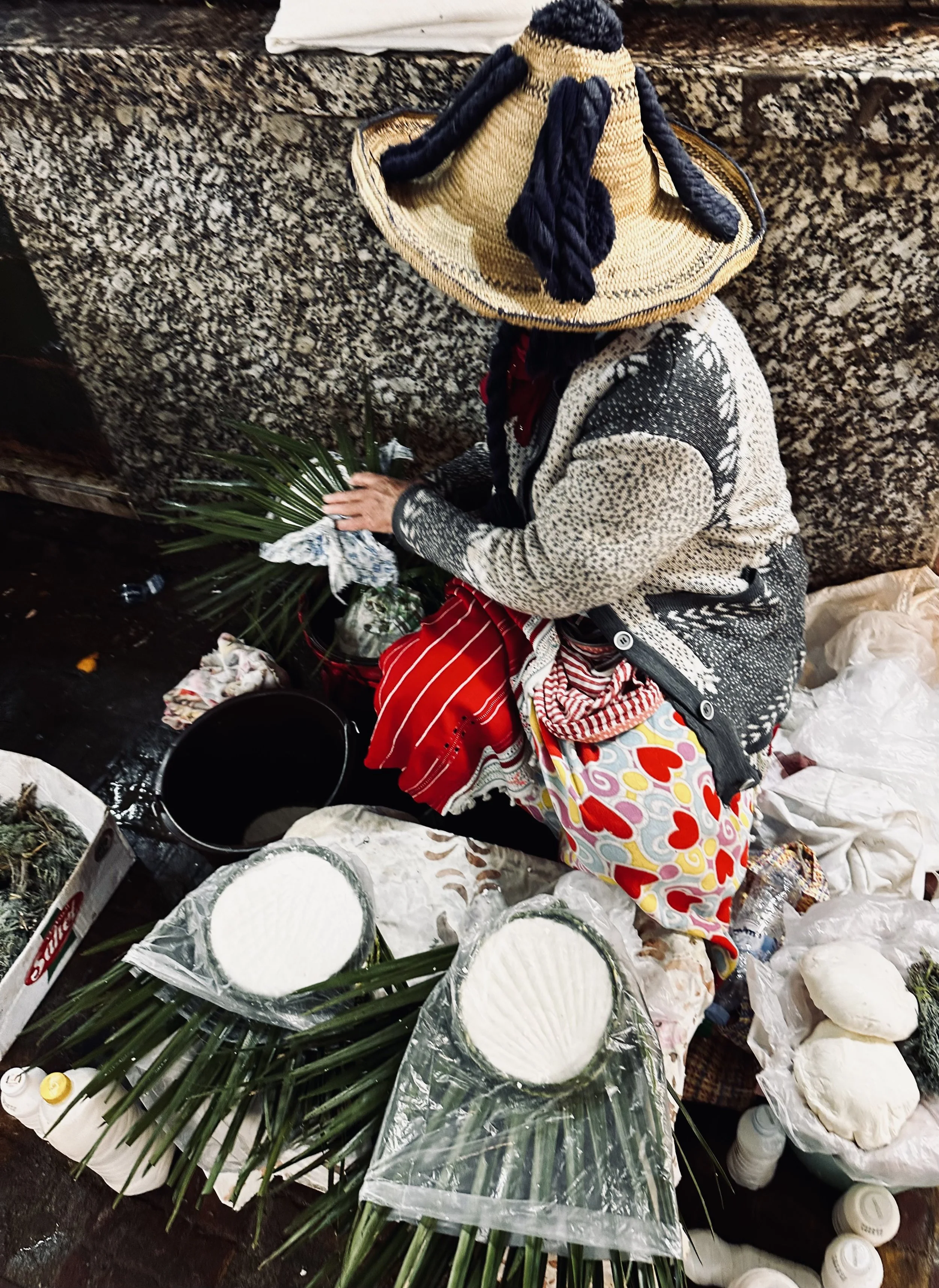 A person wearing a sombrero and patterned clothing sitting on the ground and making cheese from fresh milk. Around them are containers of cheese, palm leaves, and plastic-wrapped cheese rounds.
