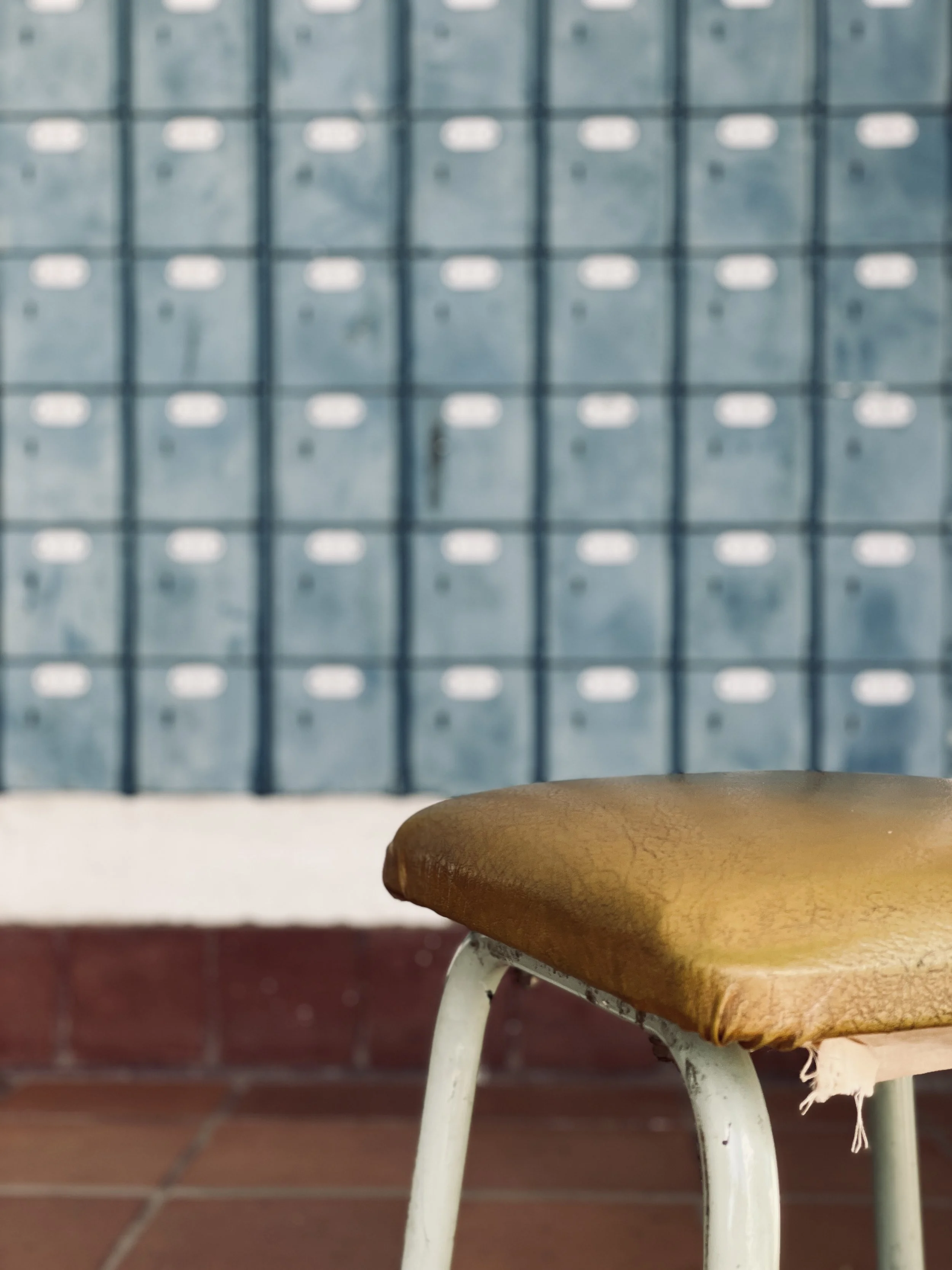 Close-up of a worn, brown vinyl seat of an old chair with a metal frame, in front of a wall of blue mailboxes or lockers.