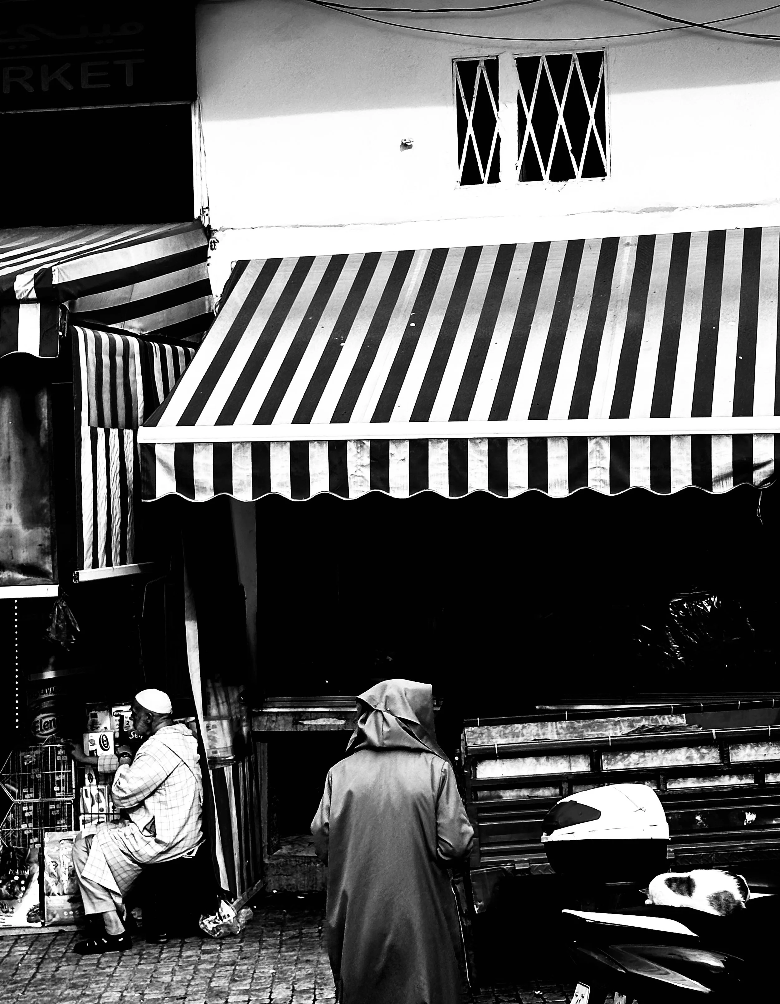 Black and white photo of a street scene with striped awnings, a man sitting on the left wearing traditional clothing and a cap, another person with a hooded jacket in the center, and a cat resting on a motorcycle on the right.